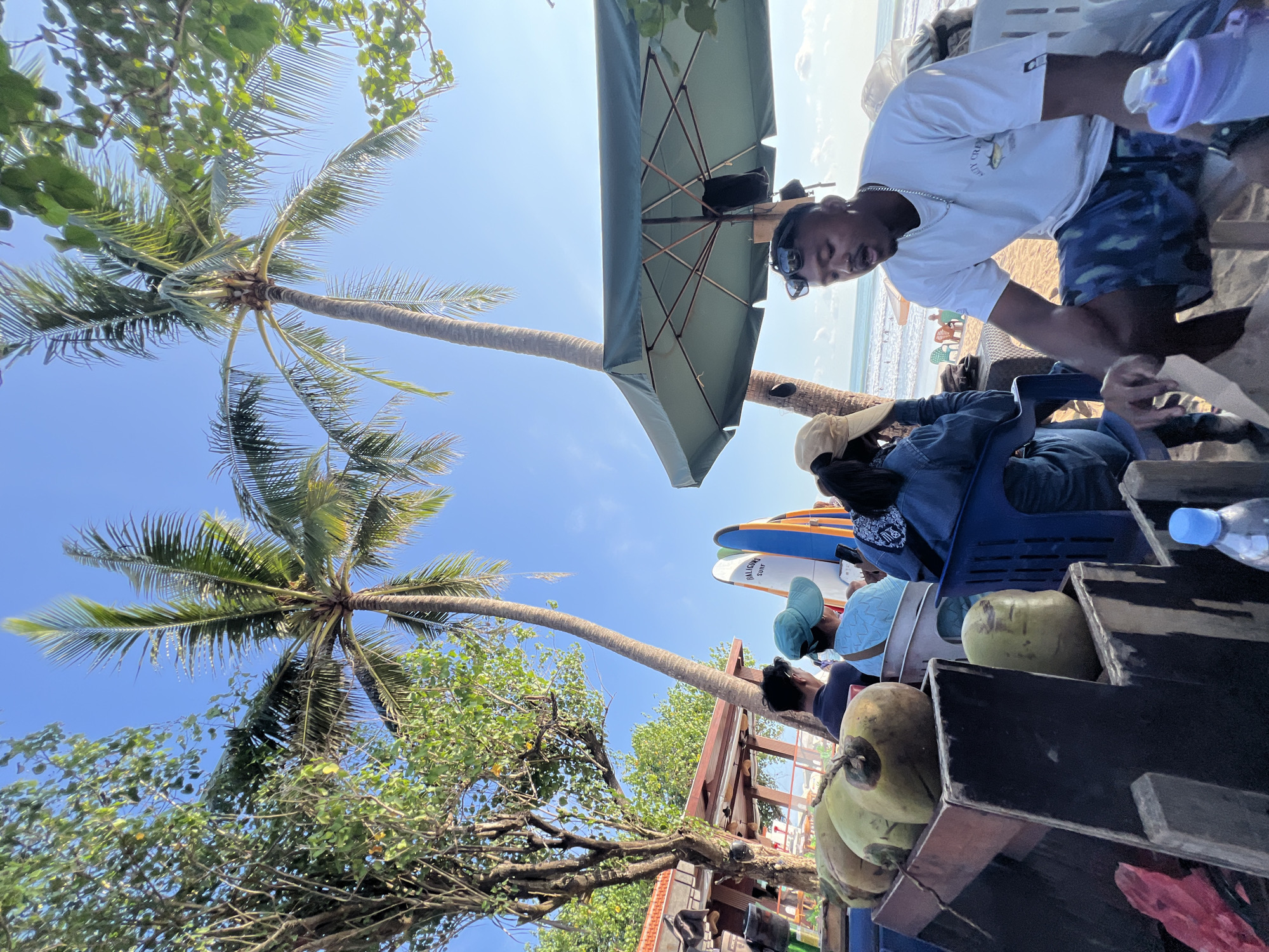 In Kuta, Indonesia, a group of people are seated at a beachside establishment. In the foreground, a man in a white t-shirt and camouflage shorts sits on a chair, looking to his left. Next to him, a person in a denim jacket and a tan hat is also seated. In the mid-ground, several coconuts are displayed in wooden crates. Behind the coconuts, a person in a light blue shirt and hat is seated. Further back, two surfboards stand upright. Palm trees and lush green foliage dominate the upper half of the image, with a clear blue sky above. The ocean is visible in the background, with some beachgoers and colorful chairs on the sand. A large green umbrella provides shade over the seating area. The scene suggests a relaxed atmosphere at a tropical beach location.