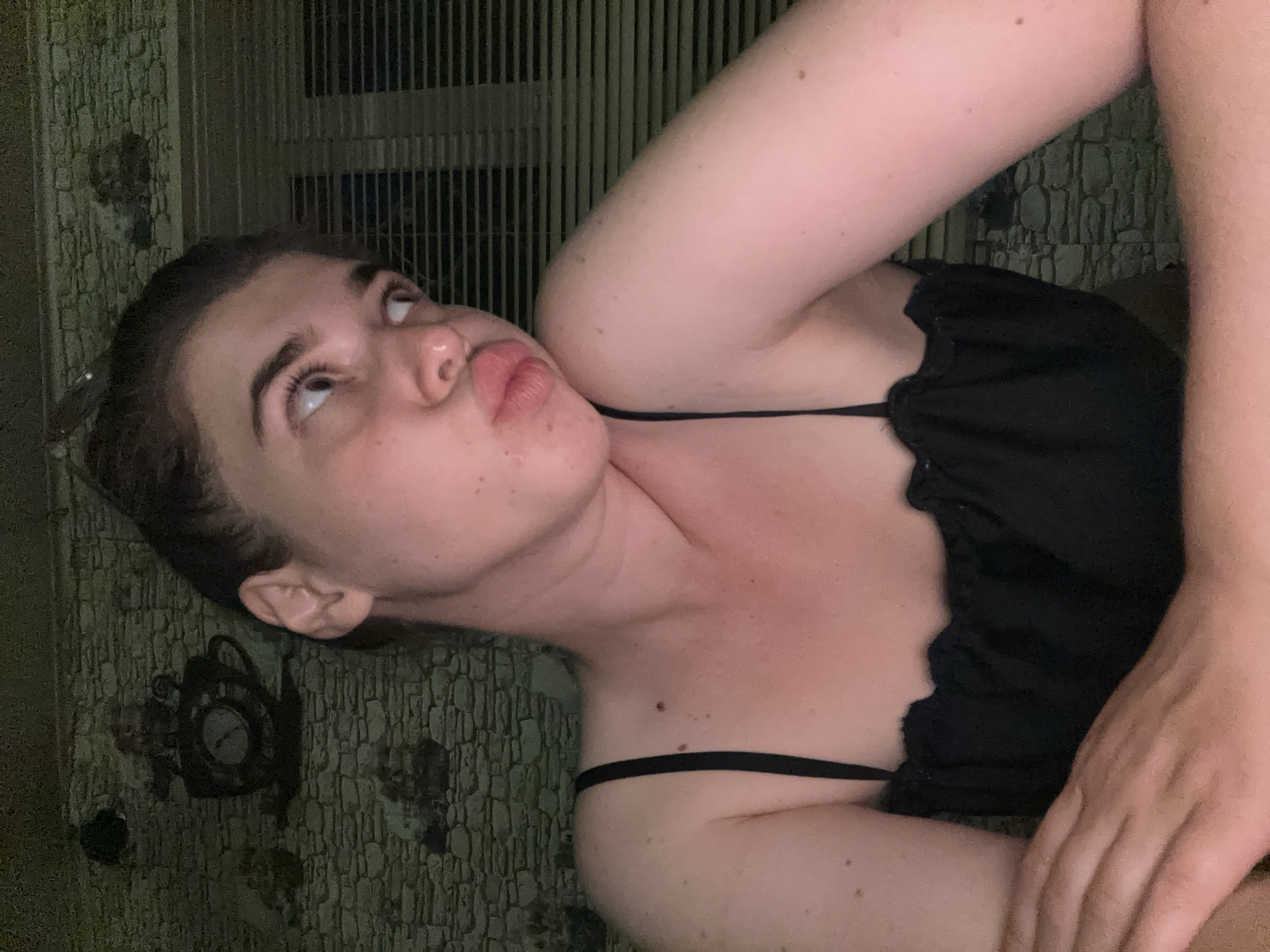 A young woman is pictured indoors, in what appears to be a home environment. She is wearing a black camisole with spaghetti straps and a ruffled neckline. Her hair is pulled back, and she is looking to her right with her lips pursed. Her arms are crossed in front of her. The background is a stone-patterned wallpaper. To the right of the woman, Venetian blinds are visible. The lighting is dim, casting a warm glow on her skin. The image does not provide information about the city of Мещёры, Russia.