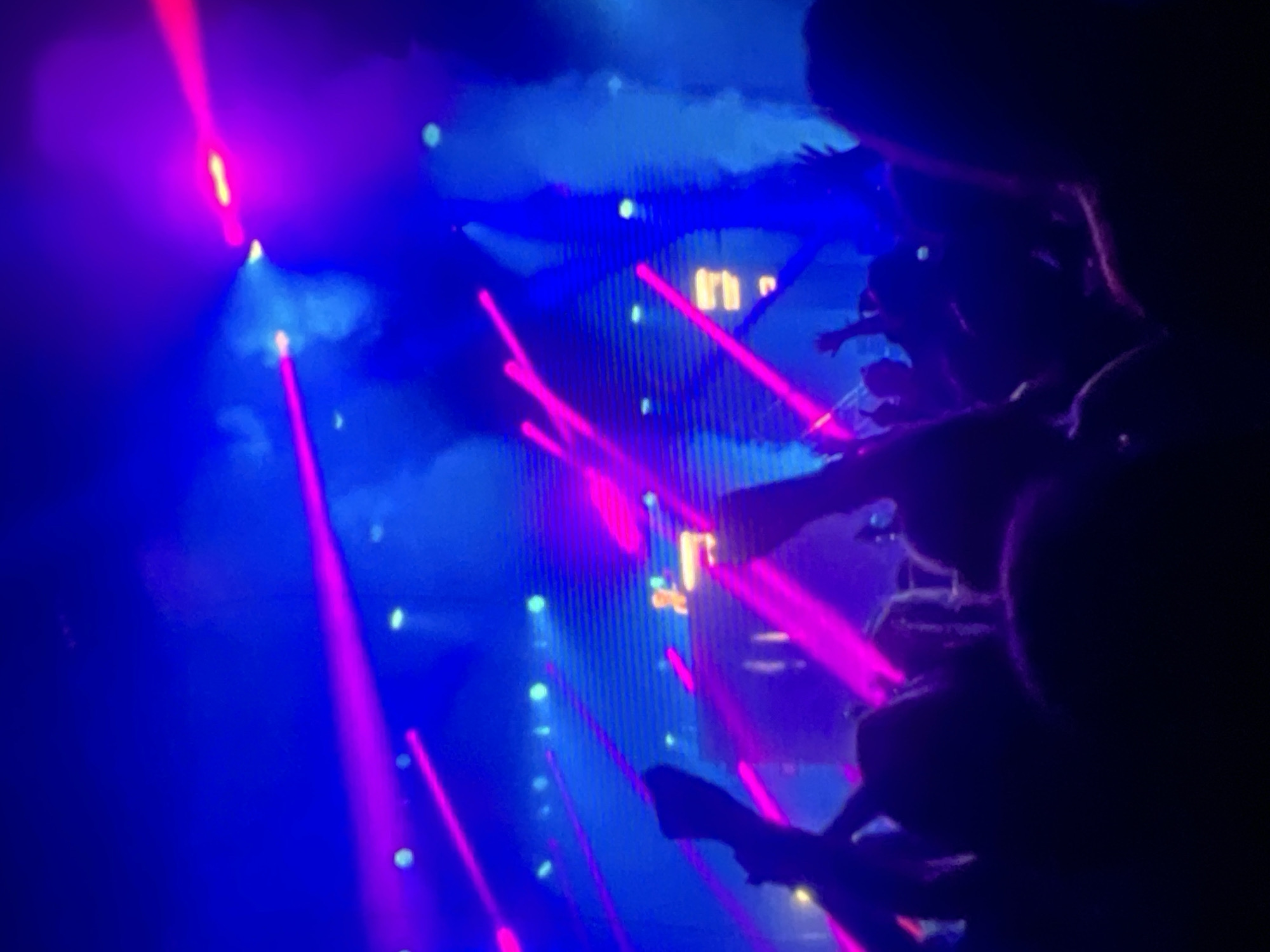 The image captures a vibrant concert or club scene in Lagos, Nigeria, bathed in a dramatic interplay of neon blue and magenta lights. The environment is filled with a thick haze, likely from fog machines, which accentuates the beams of light crisscrossing the space. In the foreground, the silhouettes of a crowd are visible, with hands raised in the air, indicating an energetic and engaged audience. Their forms are largely obscured by the low light conditions and the sheer density of the crowd.
In the mid-ground, a DJ booth or stage area is dimly illuminated, with a person, presumably the DJ, visible behind a console. The backdrop appears to be a large screen or a wall of lights, displaying a pattern of horizontal lines and illuminated panels, contributing to the overall electrifying atmosphere. The light beams, primarily in magenta, cut through the blue-toned haze, creating a dynamic and almost futuristic visual effect. The overall impression is one of a lively, high-energy entertainment event.