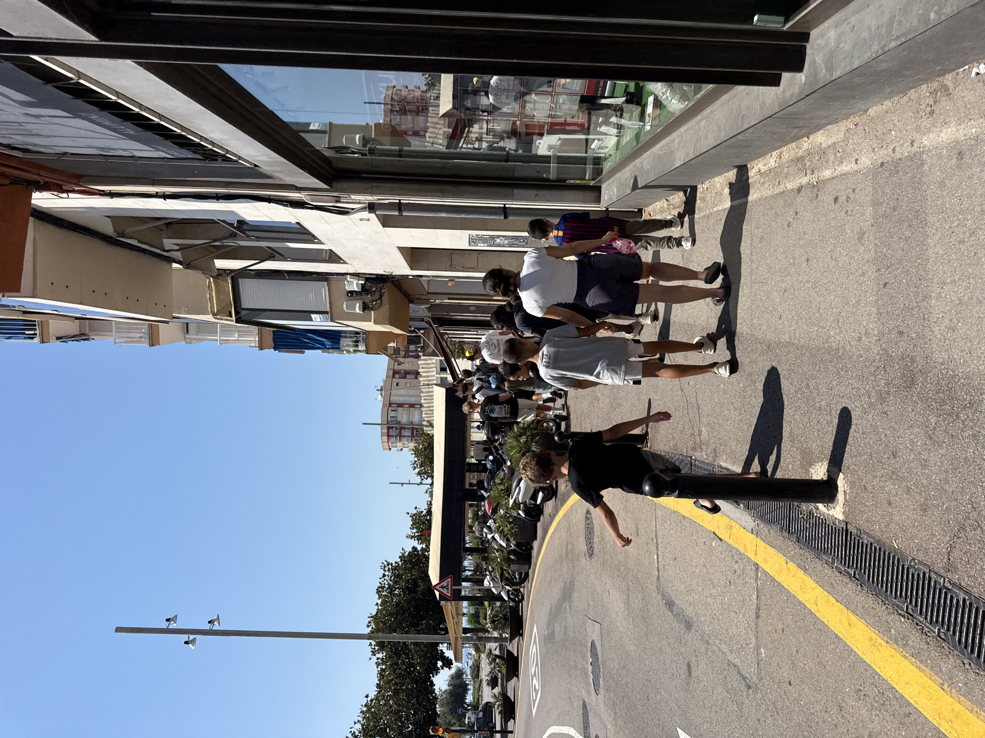 This is a daytime, eye-level, outdoor photograph captured on a clear, sunny day in Blanes, Spain. The scene depicts a street with a sidewalk adjacent to a building. On the sidewalk, a group of people, including children and adults, are walking away from the camera. A young boy in a black t-shirt and shorts is in the foreground, looking to his left as he walks. Further down the sidewalk, a woman in a white t-shirt and dark shorts walks alongside a younger boy in a grey t-shirt and shorts, and another child in a soccer jersey. To the right of the sidewalk is a storefront with large windows reflecting the street and buildings opposite. The building has multiple stories, with balconies and awnings visible. On the street side, there is a road with a yellow line marking the edge of the traffic lane, a yellow traffic light, parked motorcycles, and planters with foliage. A metal grate runs along the edge of the sidewalk next to the road. The overall atmosphere is that of a bustling, summery street scene.