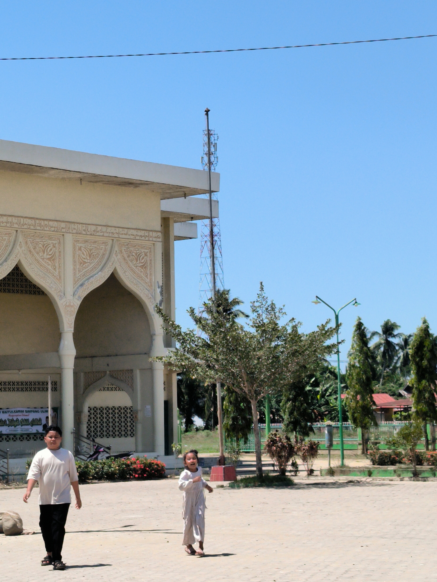 In Gampongblang, Indonesia, a building with Islamic architectural elements stands with arches and decorative inscriptions. A red and blue communication tower rises behind the building. A young boy in a white shirt and black pants walks towards the left of the frame, while a young girl in a white outfit walks to the right. The background includes green trees, shrubs, a fence, and distant buildings. The ground is paved. A banner is attached to the building, partially visible. A motorcycle is parked behind the banner. Street lights are present among the trees.
