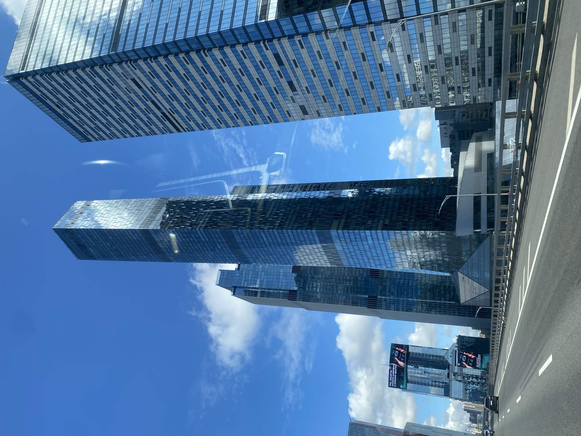 The image shows a view from a highway in Moscow, Russia, featuring a cluster of modern skyscrapers under a bright blue sky with scattered white clouds. The skyscrapers are characterized by their glass facades, reflecting the sky and surrounding buildings. On the left side of the image, a billboard is visible on one of the buildings, displaying text and images related to "ТО, ОСАГО, оплата штрафов и многое другое" (which translates to "OSAGO, payment of fines, and much more"). Several cars are driving on the highway, which has visible lane markings and guardrails. The overall scene depicts a dynamic urban landscape with prominent architectural features and a clear, sunny day.