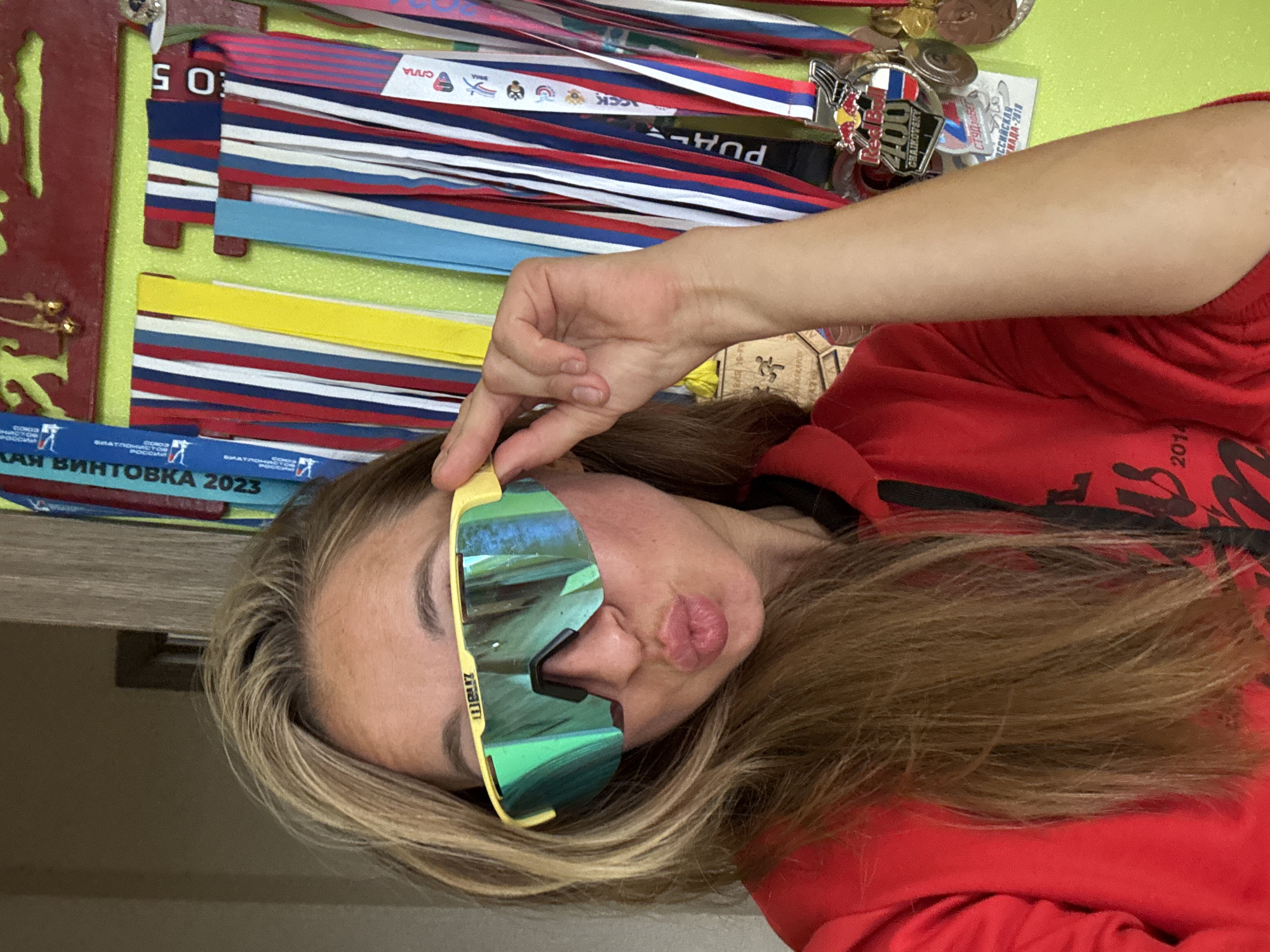 A young woman with blonde hair, wearing a red hoodie and yellow-framed sports sunglasses, puckers her lips towards the camera. Her face is turned slightly to the side, and her right hand is holding the side of her sunglasses. Behind her, a collection of medals and ribbons are displayed vertically, suggesting a sports or competition-related setting. Some of the ribbons have text and images on them, indicating participation in various events. Visible text on the ribbons and medals includes "SO", "URARCEN", "PUA", "CHAIKOVSKY", "КАЯ ВИНТОВКА 2023", "КИАДА-2018", and "2014". The medals are varied in design and size, with some featuring flags and Olympic rings. The background is a light green wall. The overall impression is of a proud athlete displaying her achievements.