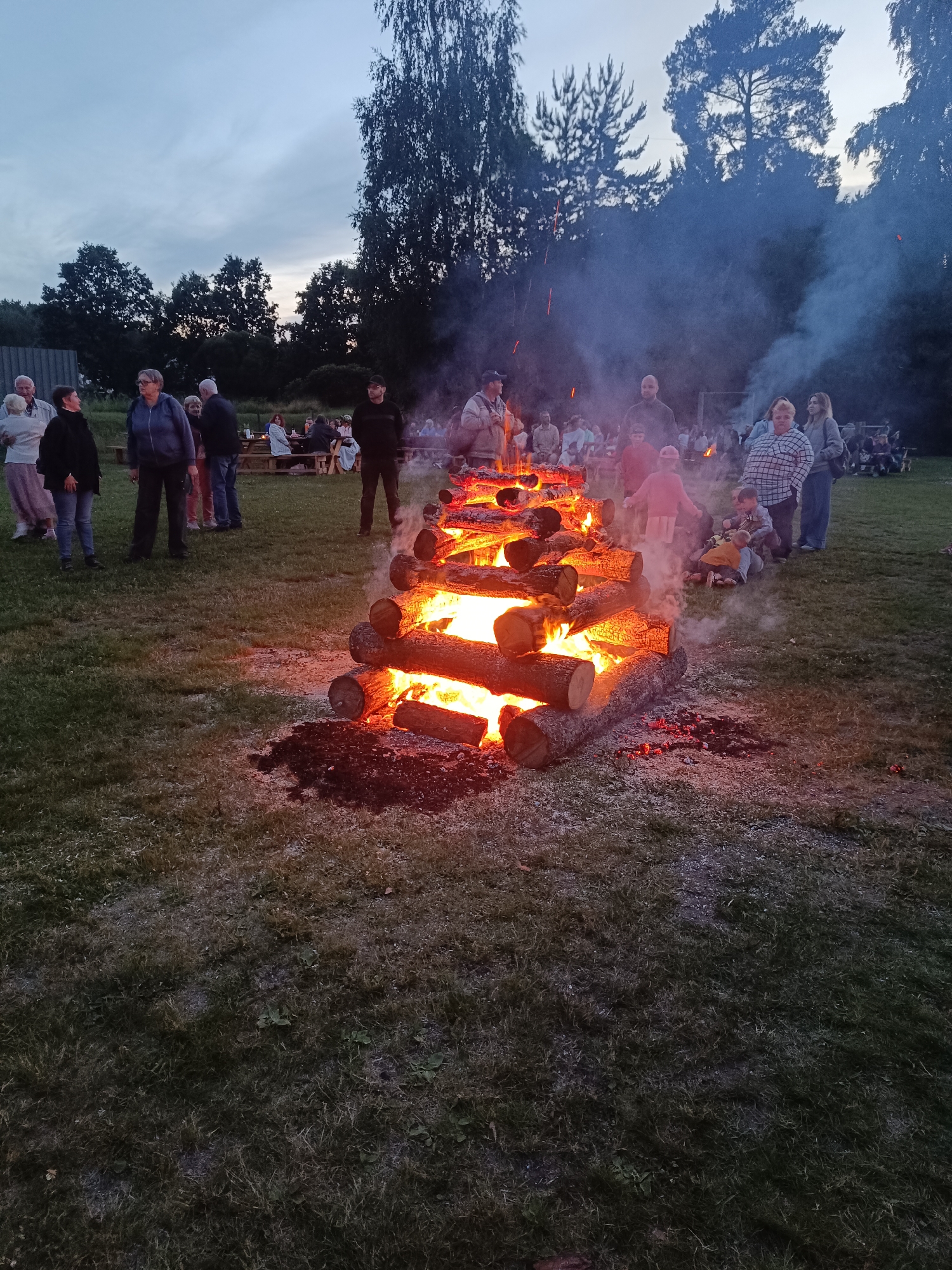 The image captures a midsummer celebration, likely Jāņi, in Carnikava, Latvia. The scene is set outdoors in a grassy field with trees forming a dark silhouette against the twilight sky. A large bonfire made of stacked logs is the central focus, flames licking upwards and smoke billowing into the air. The fire is actively burning, with glowing embers and sparks visible.
Around the bonfire, a crowd of people are gathered, some standing and watching the spectacle, while others are seated on the grass or at picnic tables in the background. Children are also present, with some sitting close to the fire. The mood appears relaxed and communal, with people enjoying the evening outdoors. The time of day is clearly evening, indicated by the dim, overcast sky and the artificial light from the bonfire. The weather appears to be calm, with no visible signs of rain or strong wind.
The people in the foreground are dressed in casual attire, suggesting a relaxed, informal gathering. The overall atmosphere is festive and social, typical of a summer celebration in Latvia. There is no visible text in the image.