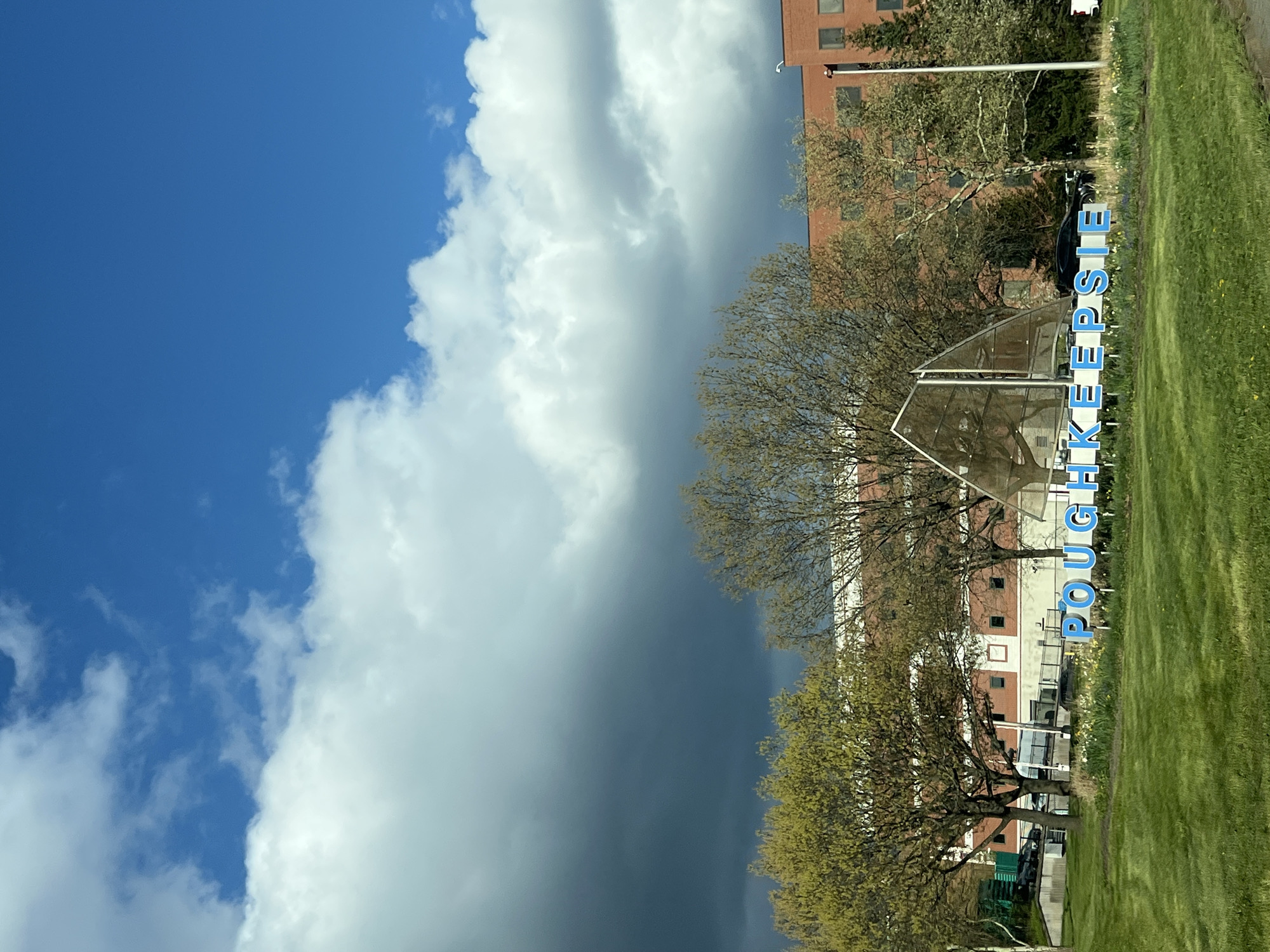 The image shows a view in the City of Poughkeepsie, New York. The sky is divided, with a clear blue sky with scattered white clouds on the left side, and a darker, ominous-looking storm cloud dominating the right side of the frame.
In the middle ground, there are trees with emerging green leaves, partially obscuring a brick building with windows. A metal sculpture, possibly a sign indicating the location, is positioned in front of the trees.
In the foreground, there's a patch of green grass, possibly a lawn or park area, with the word "POUGHKEEPSIE" spelled out vertically in large, blue letters, likely part of a sign or landmark. The letters are oriented sideways in relation to the image's current orientation.
The scene appears to be a moment captured during a weather transition, with a clear sky juxtaposed against an approaching storm front. The image appears to be rotated ninety degrees counter-clockwise, distorting the typical perspective.