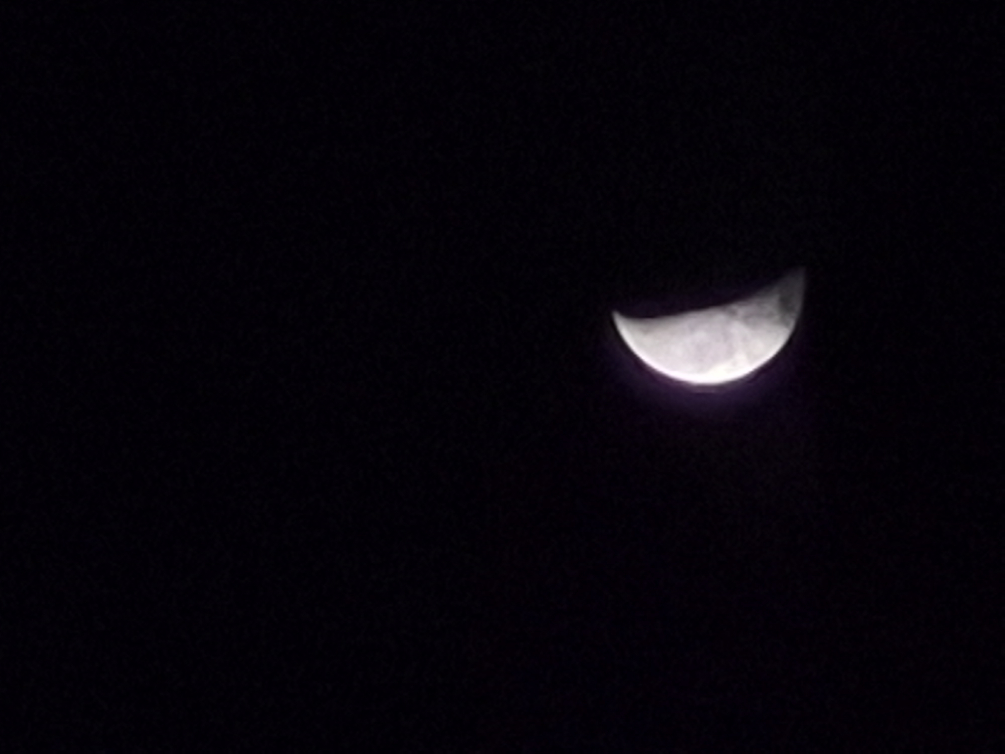 The image is a close-up, vertical shot of a crescent moon against a black sky. The moon is positioned in the lower right quadrant of the frame. Its illuminated portion is a bright, pale white with subtle gray markings suggesting craters or surface features. The dark portion of the moon is indistinguishable from the surrounding blackness of space. The image provides no information about a specific location in the city of Sooblase, Estonia, or any context related to human presence, actions, or objects other than the celestial body.