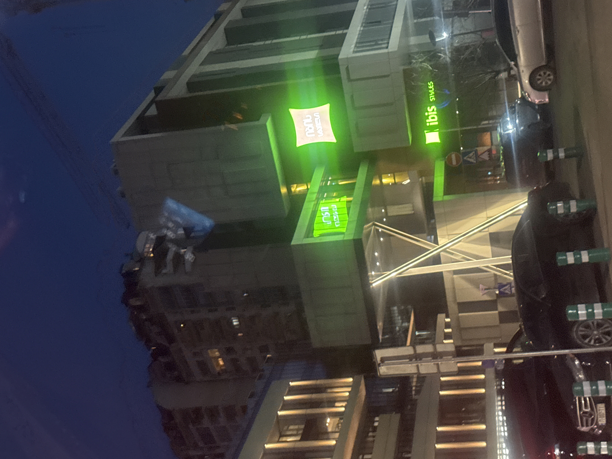 An exterior, nighttime, low-angle shot of a building in the city of Tbilisi, Georgia. The building features a well-lit "ibis Styles" sign. Adjacent to the hotel is a multi-story structure with illuminated horizontal bands. A dark-colored vehicle is parked on the street alongside the building, near green-striped bollards. A bright street lamp lights the scene. A building is visible in the distance.