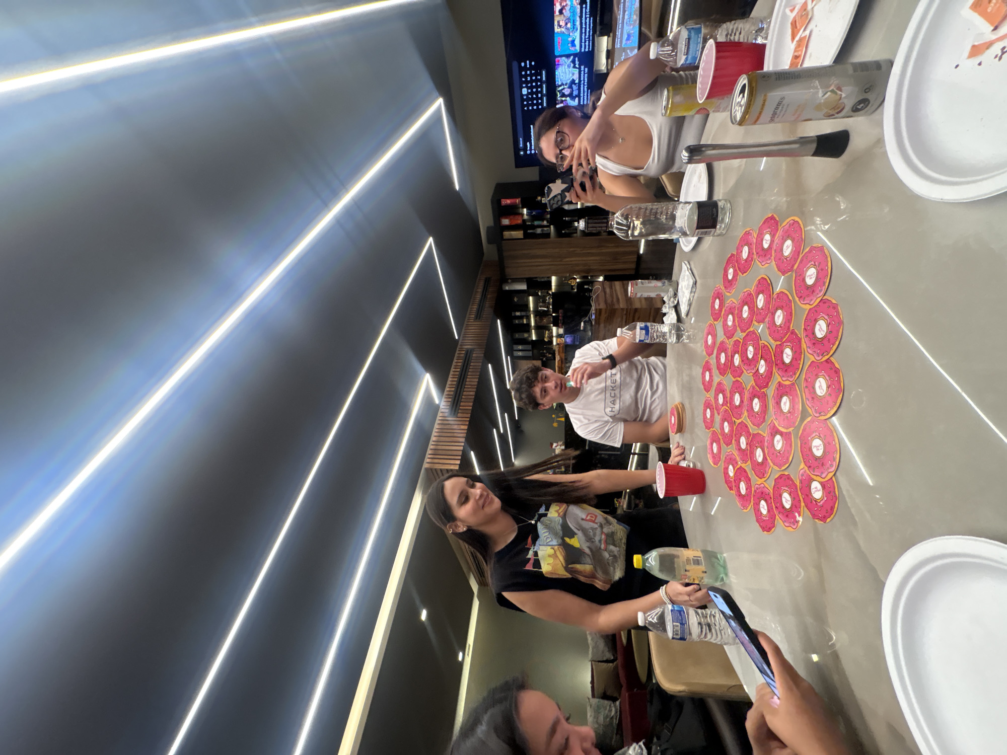 In San Bernardino Tlaxcalancingo, Mexico, a group of people are gathered around a table. On the table are stacks of donut-shaped game pieces, plates, cans, water bottles, and red plastic cups. The people are interacting with each other and using their phones. The environment appears to be a modern interior with linear light fixtures on the ceiling and a bar in the background.