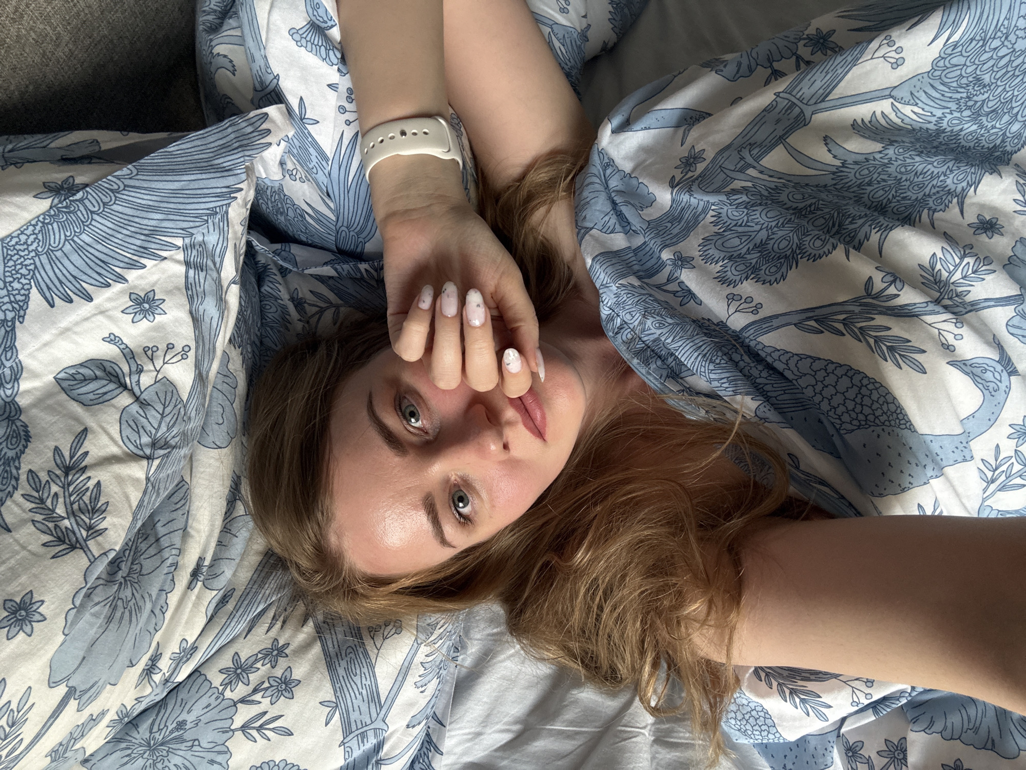 A woman with light brown hair and fair skin is lying in bed on a patterned blue and white duvet cover. She is looking directly at the camera with a slight smile. Her hands are raised in front of her face, one adorned with a white wristwatch. Her fingernails are painted with a light design. The bedding features a repeating pattern of blue and white motifs depicting birds, flowers, and branches. The location and time cannot be determined.