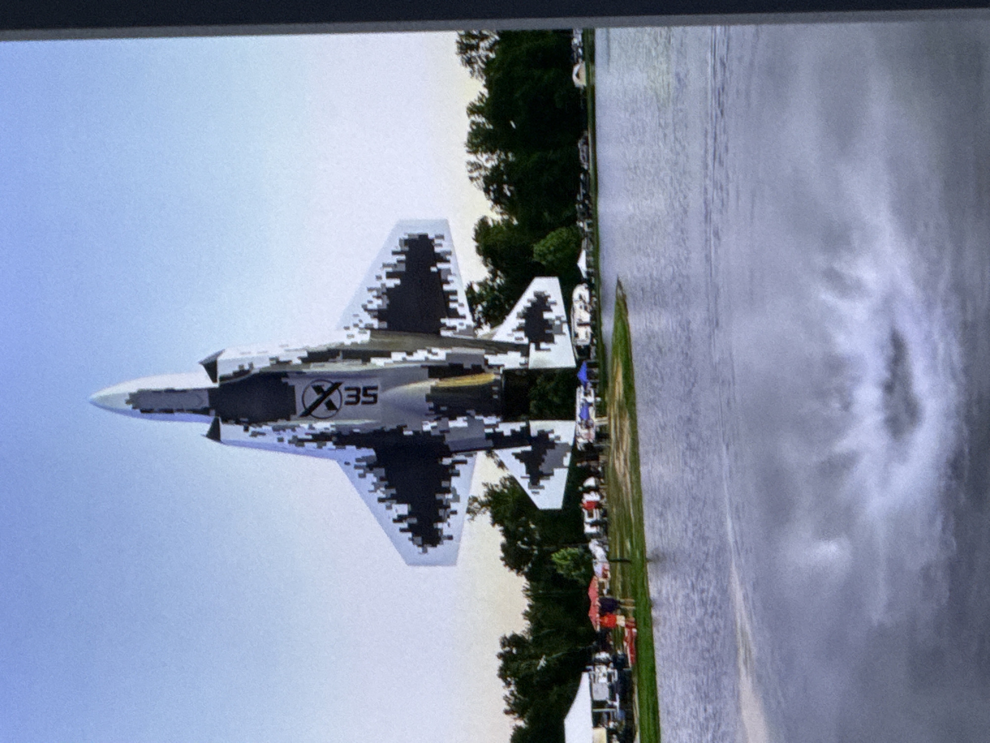 An aerial photograph captures a remotely operated model jet, designed to resemble an F-35 fighter jet, in mid-flight above a body of water. The model, adorned with a pixelated camouflage pattern and the marking "SEX35", is positioned prominently in the upper center of the frame. Below the jet, a shoreline is visible, lined with trees and a grassy area where several tents, people, and what appear to be parked vehicles are present. The water's surface reflects the sky and the jet, with a distinct splash or disturbance in the water directly beneath the aircraft, suggesting it may be performing a water maneuver or has just touched down. The sky is clear and pale blue. The specific location in the city of Cahul, Moldova, cannot be determined from the visual information provided in the image.