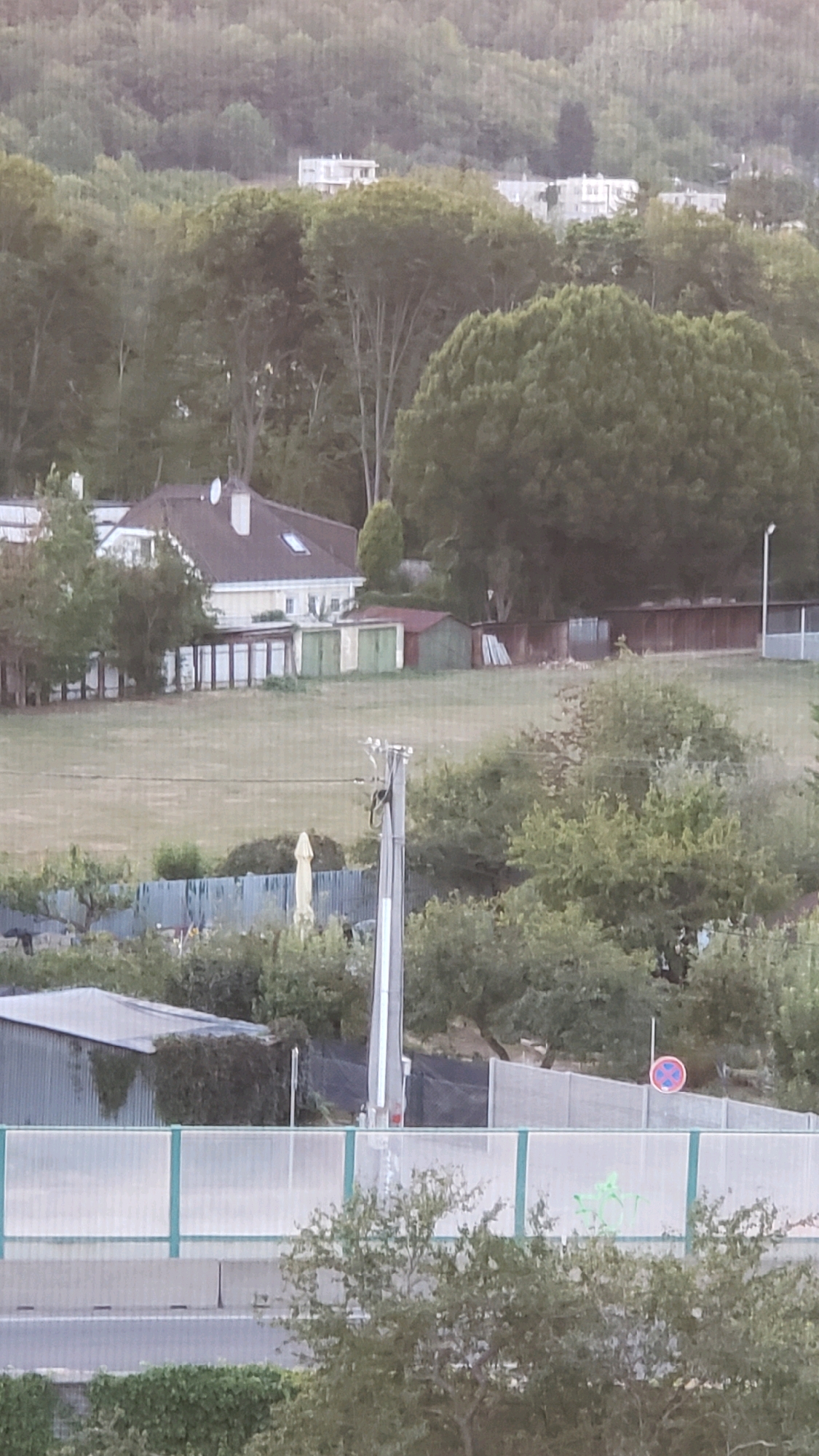 The image captures a view of a residential area with trees and buildings, likely in the city of Prešov, Slovakia. In the foreground, a road is visible, bordered by a modern sound barrier. Behind the barrier, a large expanse of green grass is seen, with a house with a red roof and several outbuildings, including garages, situated on the left side of the frame. Further in the background, dense foliage and trees dominate the landscape, with some multi-story buildings peeking through the trees on a distant hillside. The overall impression is of a suburban or semi-rural setting, possibly on the outskirts of the city.
