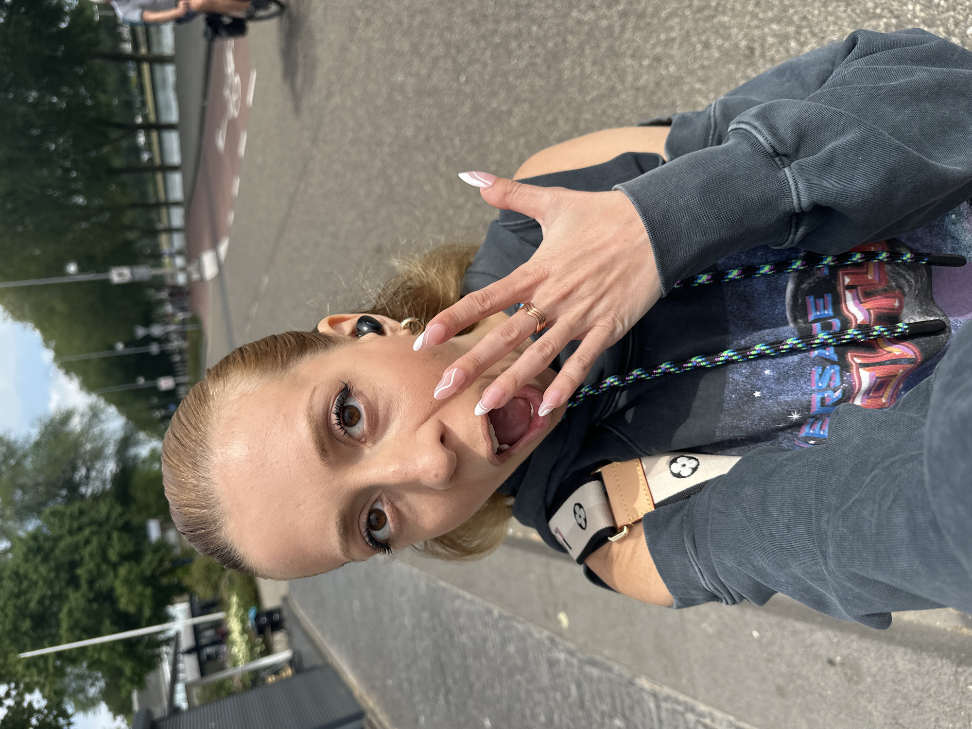 A woman with blonde hair pulled back, brown eyes, and open mouth, is captured in a selfie outdoors. She is wearing a dark gray hoodie with cut-out shoulders and a graphic print of a galaxy with text. On her left ear are black wireless earbuds, and on her right hand, she wears a gold ring and has long, white French manicured nails. A light brown strap with a white and black logo runs across her shoulder. The background shows a paved walkway with trees lining a road. Beyond the road, there is a body of water, and in the distance, more trees and buildings are visible. The scene appears to be a street in Amsterdam, Netherlands, with a bicycle path visible on the road. A person on a bicycle is also visible in the background.