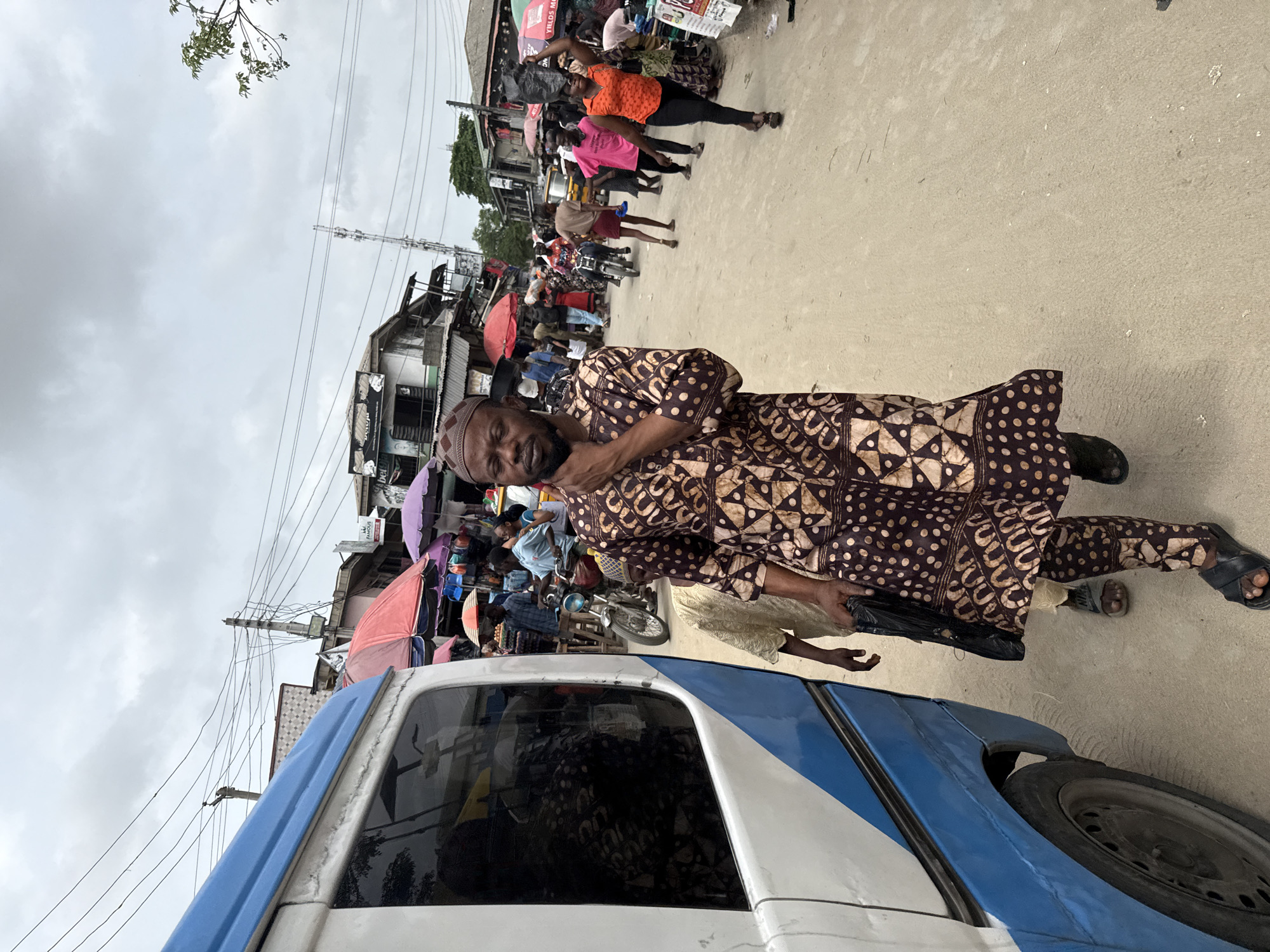 This media file depicts a vibrant and bustling street scene in Ado, Nigeria, under an overcast, cloudy sky. The setting is an active market or commercial area with an unpaved street, possibly dirt or rough concrete.
In the prominent foreground, a man of West African descent looks directly at the camera with a neutral to slightly serious expression. He is dressed in traditional brown attire featuring distinct patterned tunic and matching pants, and wears a brown cap. He stands near a partially visible blue and white van or minibus, holding a dark object, possibly a bag, in his left hand.
The mid-ground and background are teeming with people, indicating a busy daily life scenario. Individuals in various casual and traditional clothing are seen walking, standing, and engaging in market activities. A woman in an orange top and black pants is noticeable, carrying a large black bag balanced on her head. Other market-goers and vendors are visible amidst numerous colorful market umbrellas (red, purple, orange) that shade stalls selling various goods. A motorcycle or moped is also parked among the crowd.
The street is lined with simple, multi-story buildings, some appearing aged. Overhead, a complex network of utility poles and wires crisscrosses the sky. Sparse green foliage is visible against the muted light of the cloudy day. Visible text on partially obscured signs includes fragments like "YIELDS MOR..." and possibly "FAMOUS" on market stalls. The overall atmosphere is one of lively commerce and community interaction during daytime.