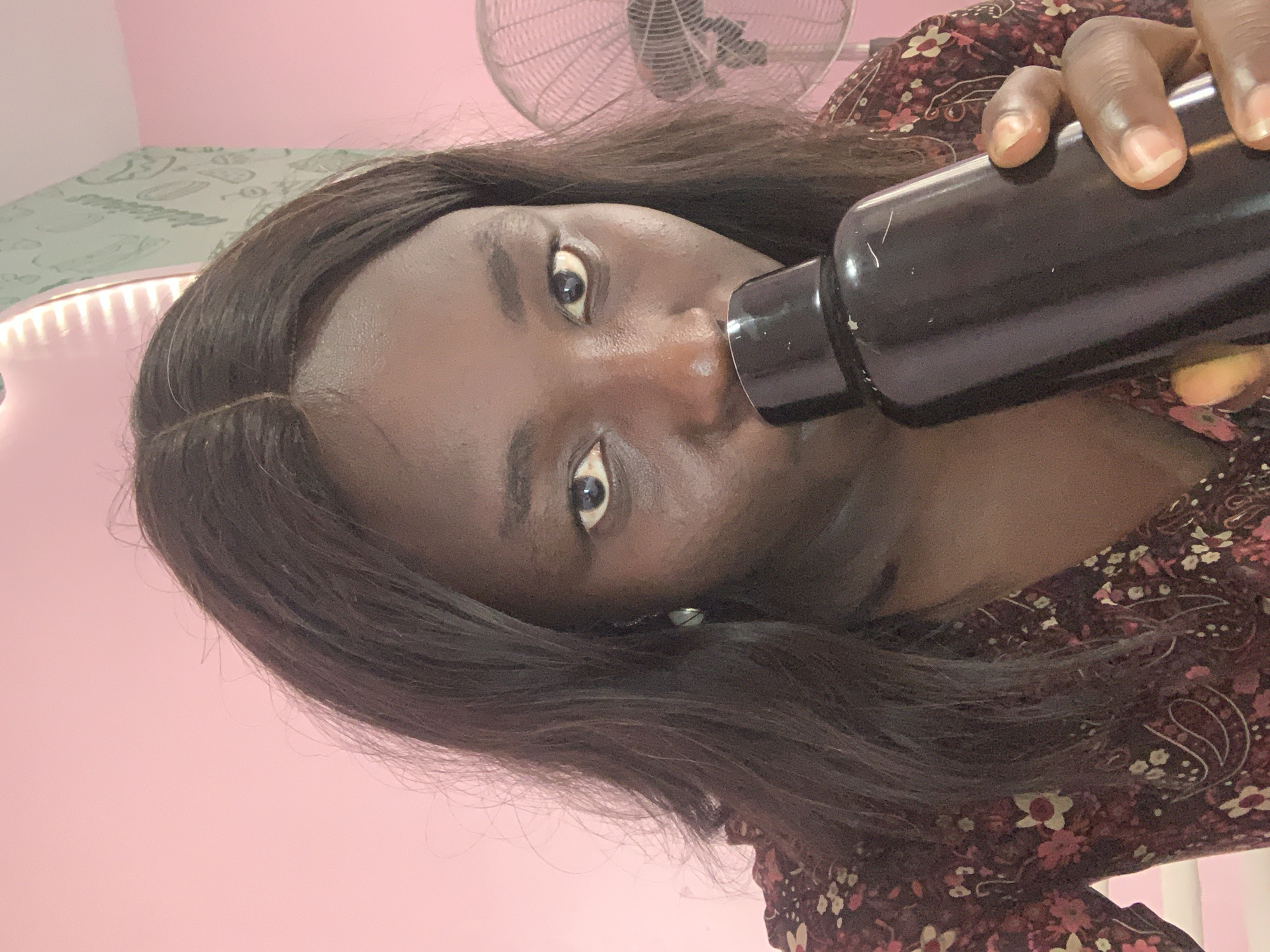 In a close-up, head-and-shoulders selfie, a young Black woman with dark, wavy hair is shown from the chest up. She is holding a dark-colored water bottle to her lips with her right hand. Her eyes are open and looking directly at the camera. She is wearing a patterned top with a floral design in shades of red, pink, and white. The background consists of a pink wall and a partially visible fan on the right side. The lighting suggests an indoor environment, possibly a bedroom. The scene does not contain any specific indicators to place it in Jos, Nigeria.