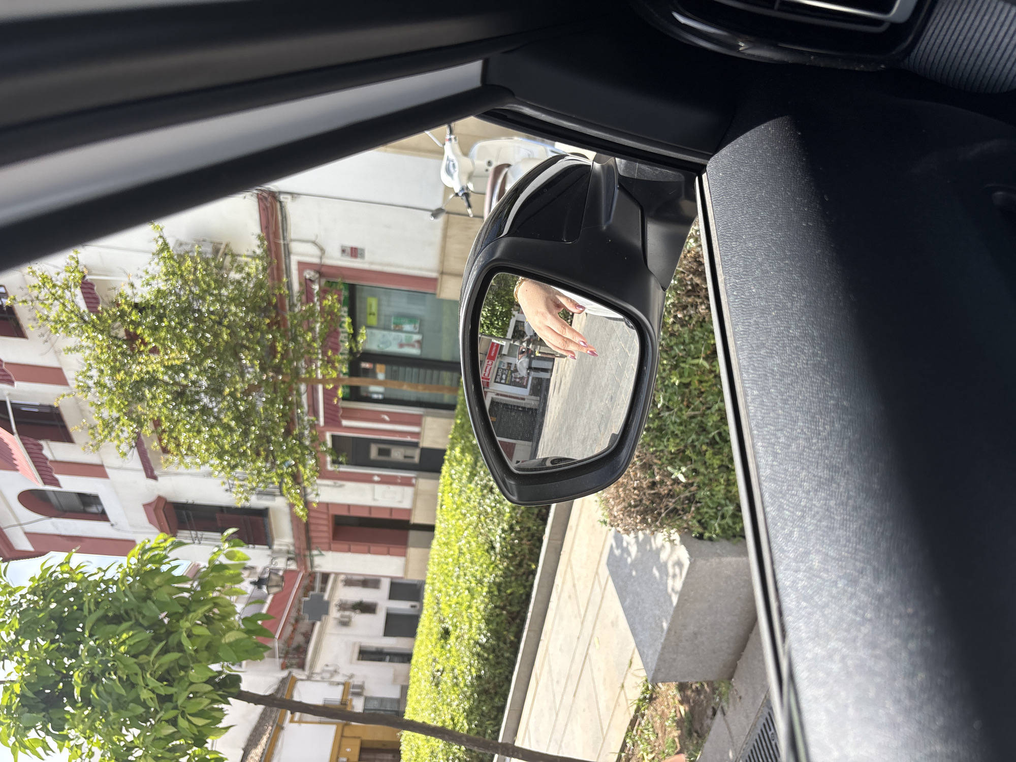 The image captures a sunlit view from the interior of a car, looking out through the passenger side window onto a street in Córdoba, Spain. The original image appears to be rotated 90 degrees clockwise, indicating it was likely taken with the camera held sideways.
In the immediate foreground, dark gray car interior elements are visible, including a textured door panel and the black casing of a side mirror. The side mirror prominently reflects a person's hand with manicured, painted nails (appearing a pinkish-purple hue), suggesting a passenger is present.
Beyond the car's interior, the scene reveals an urban environment bathed in bright daylight, casting clear shadows. Lush green foliage from trees and bushes lines the street, with some branches extending into the frame from the top and bottom. In the background, multi-story buildings with white facades and red or terracotta-colored window frames and awnings line the street, characteristic of Spanish architecture. A light-colored scooter or moped is partially visible, parked on the street. A concrete sidewalk with visible seams runs alongside the buildings.
The time of day is clearly daytime, given the bright light and distinct shadows, suggesting sunny weather. Although some text is visible in reflections and on distant storefronts, it is too blurry and small to be deciphered. The overall impression is a vibrant, typical street scene in a Southern European city.