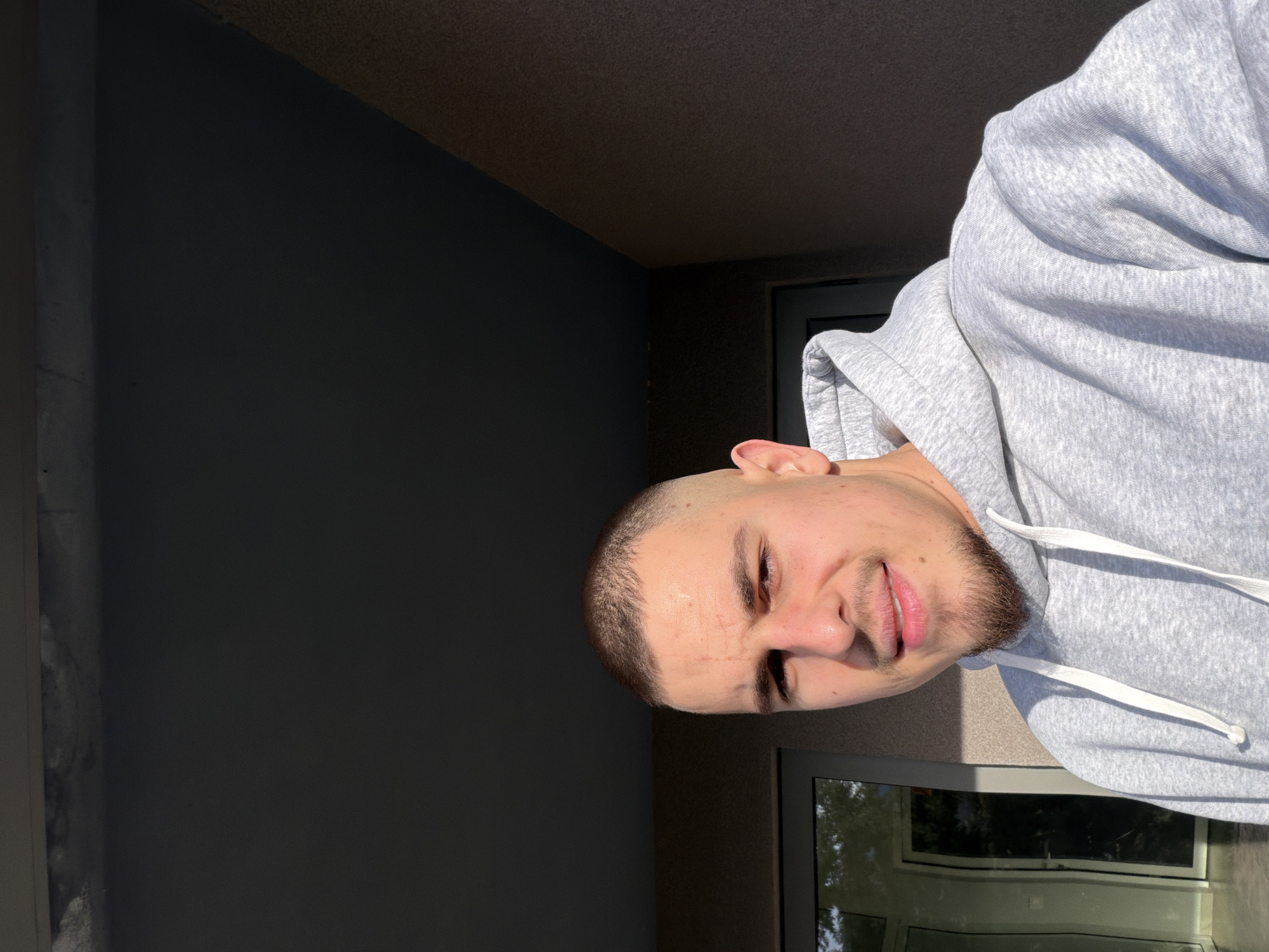 A man is posing for a selfie in a residential setting. He is wearing a light grey hoodie and has a short haircut and a trimmed beard. The background shows the exterior of a building, including a dark grey wall and a window. The reflection in the window suggests outdoor greenery. The setting appears to be on a balcony or near a window in Parvenets, Bulgaria.