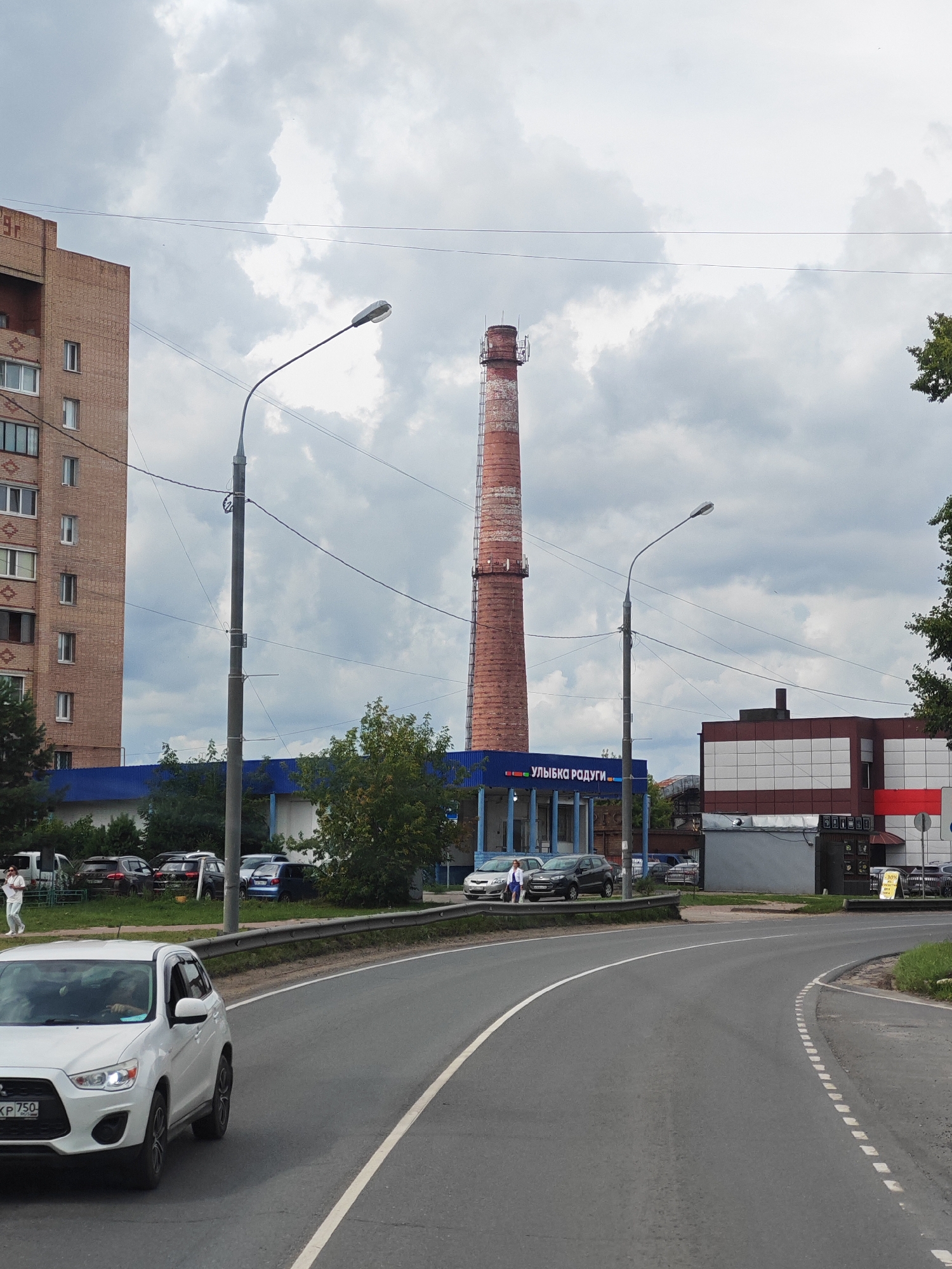 This is a wide shot of a street scene in Likino-Dulyovo, Russia, on a cloudy day. In the foreground, a white Mitsubishi ASX is driving on a gray asphalt road. The car has a license plate that reads "XK 750". Behind the car, the road curves to the right.
Further down the road, there are several parked cars and a blue building with a sign that reads "УЛЫБКА РАДУГИ" (YLYBKA RADUGI), which translates to "Smile of the Rainbow". To the left of the blue building is a tall, red brick smokestack, indicating an industrial area. To the right of the blue building is a brown and white building with a red stripe, possibly a commercial establishment.
On the left side of the image, a multi-story brick apartment building is visible. A lamppost stands on the right side of the road, and power lines crisscross the sky. A few people are visible walking on the sidewalk near the blue building. The overall environment appears to be a mix of residential, commercial, and industrial elements within a city.