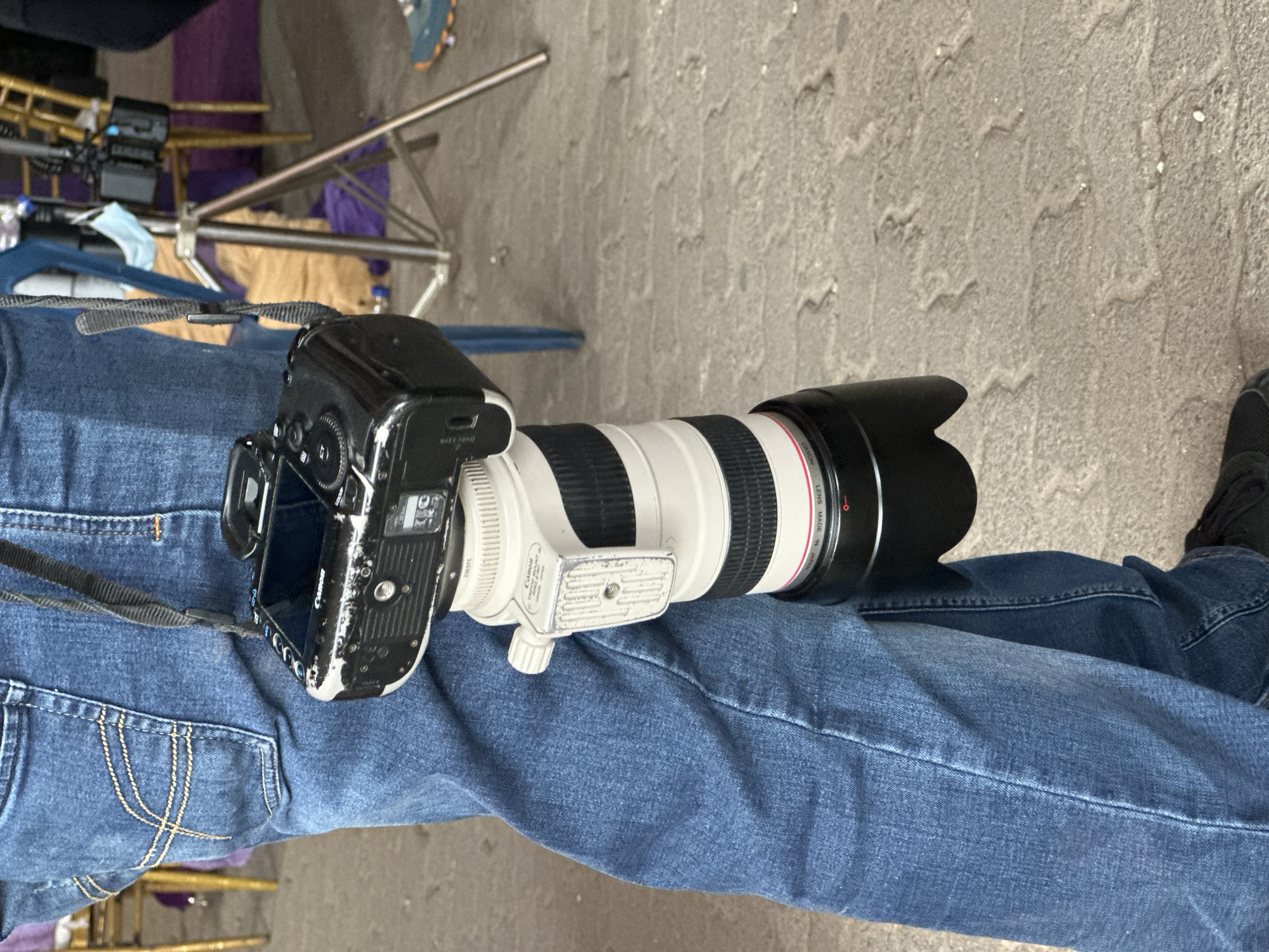The image shows a Canon DSLR camera with a long telephoto lens resting on a person's blue denim jeans. The camera has a neck strap. The background is a concrete surface, and partially visible is film or photography equipment. There is no information given indicating that the image was taken in Lagos, Nigeria. Therefore, I cannot describe the location.