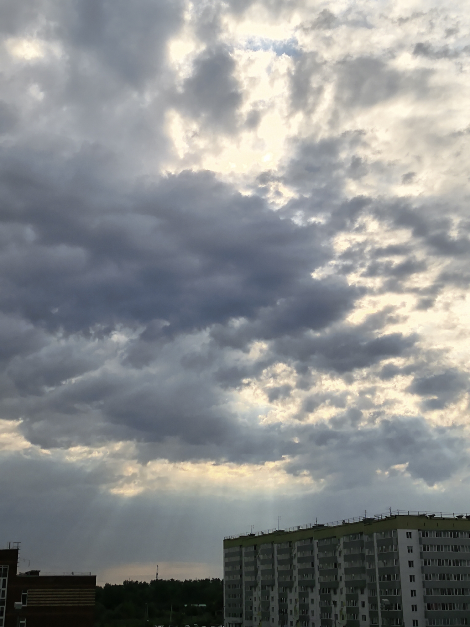 The image shows a cloudy sky over a residential area in Фролы, Russia. The sky is filled with grey and white clouds, with some patches of light breaking through. Below the clouds, there are several buildings, including a tall apartment building with many windows. There is also a smaller building with a dark roof visible to the left, and trees in the background. The overall impression is of a cloudy day in an urban environment.