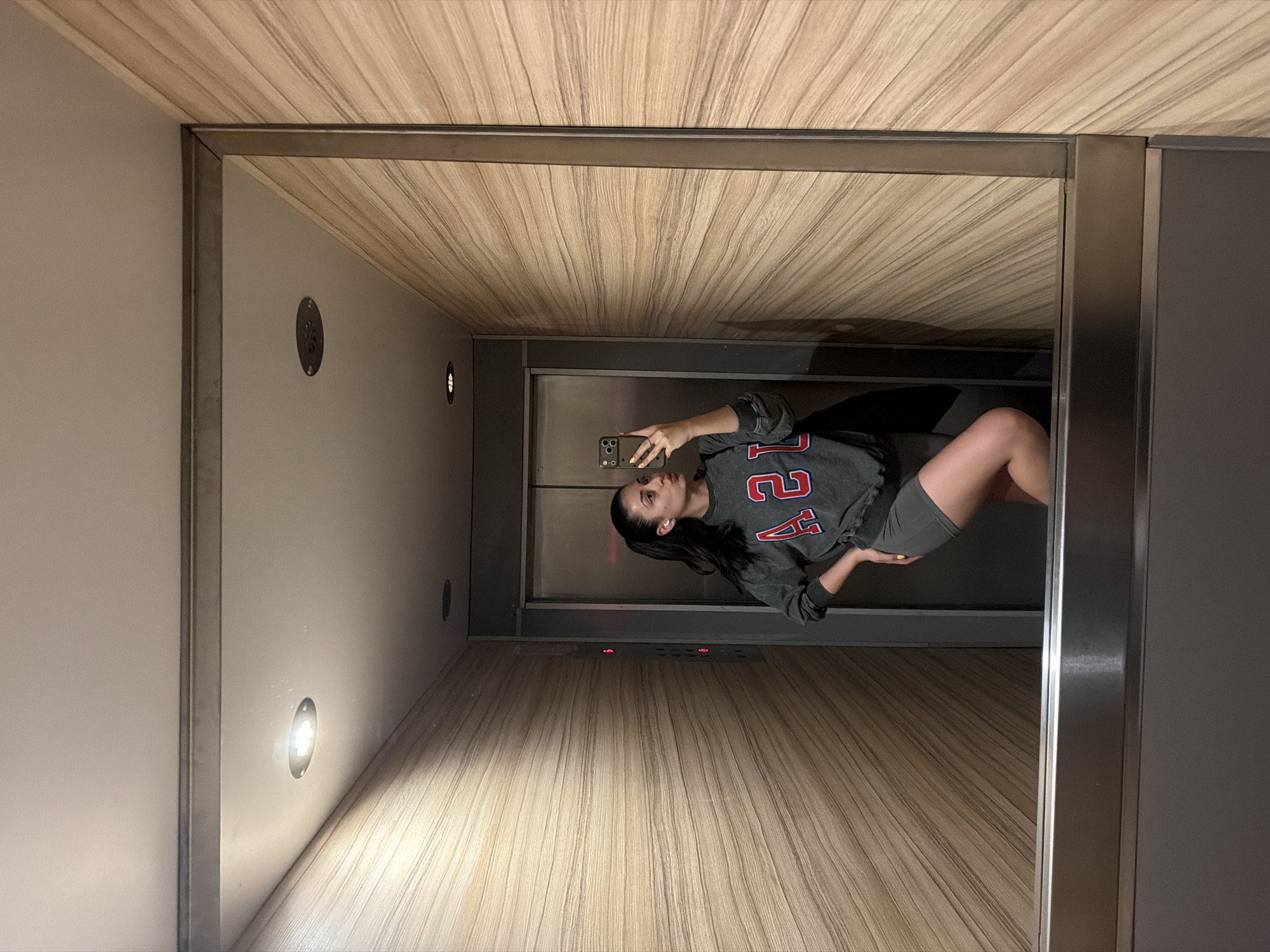 A person is taking a selfie in an elevator. The interior of the elevator is modern, with wood-paneled walls and ceiling, and recessed lighting. The person is wearing a grey sweatshirt with red lettering and grey shorts. The elevator doors are metal. The exact location in Sofia, Bulgaria, cannot be determined from the image.