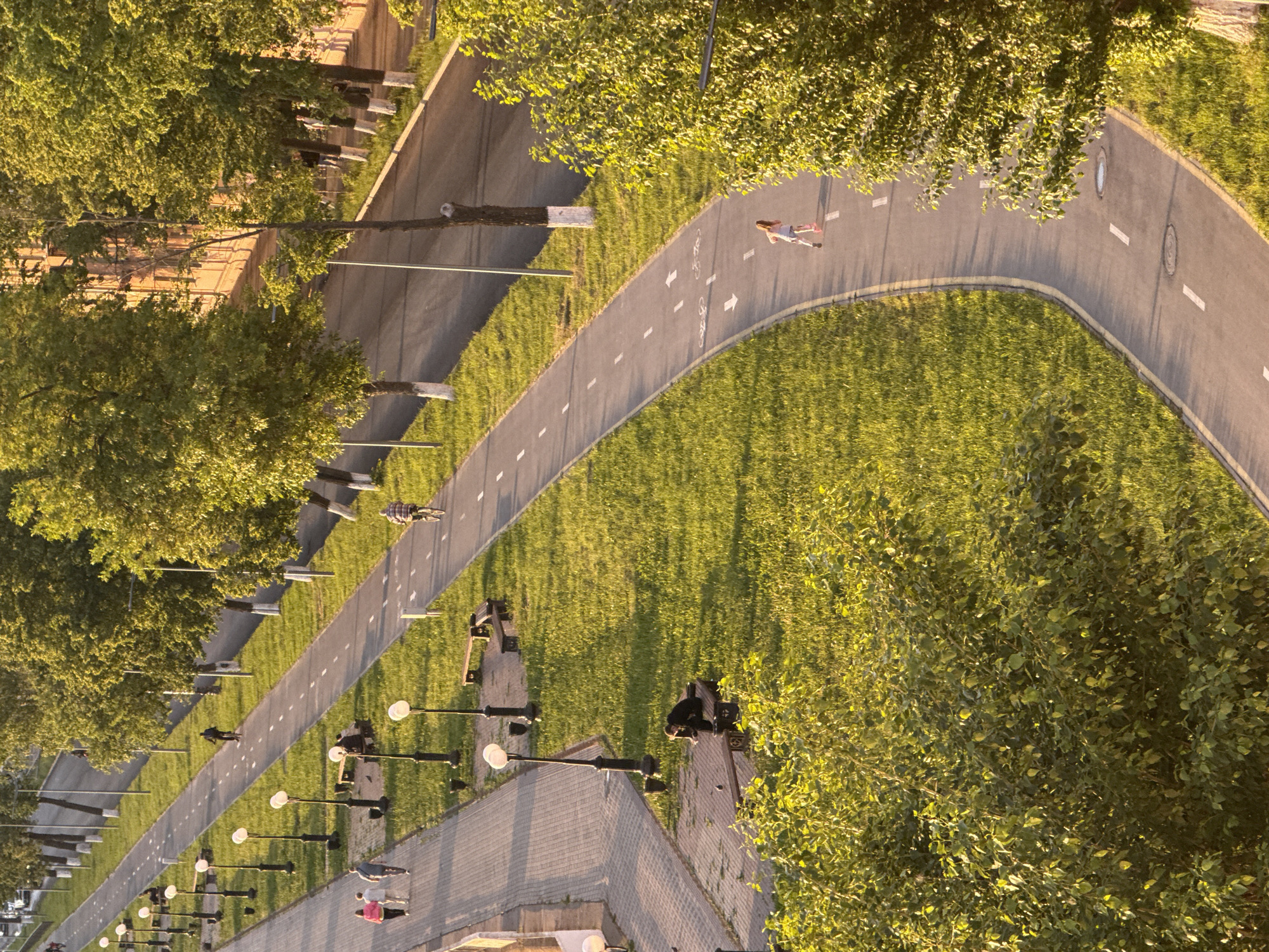 An aerial view shows a park scene in Votkinsk, Russia. A winding asphalt pathway stretches through a green space, marked with painted bicycle symbols and directional arrows. A person is running on the curved section of the path, while another individual is riding a bicycle on a straight segment further down. Rows of evenly spaced lampposts line the walkway adjacent to the path. The park also features trees and benches, with some individuals seated on the benches. The overall impression is a well-maintained park environment with recreational areas for walking, running, and cycling.