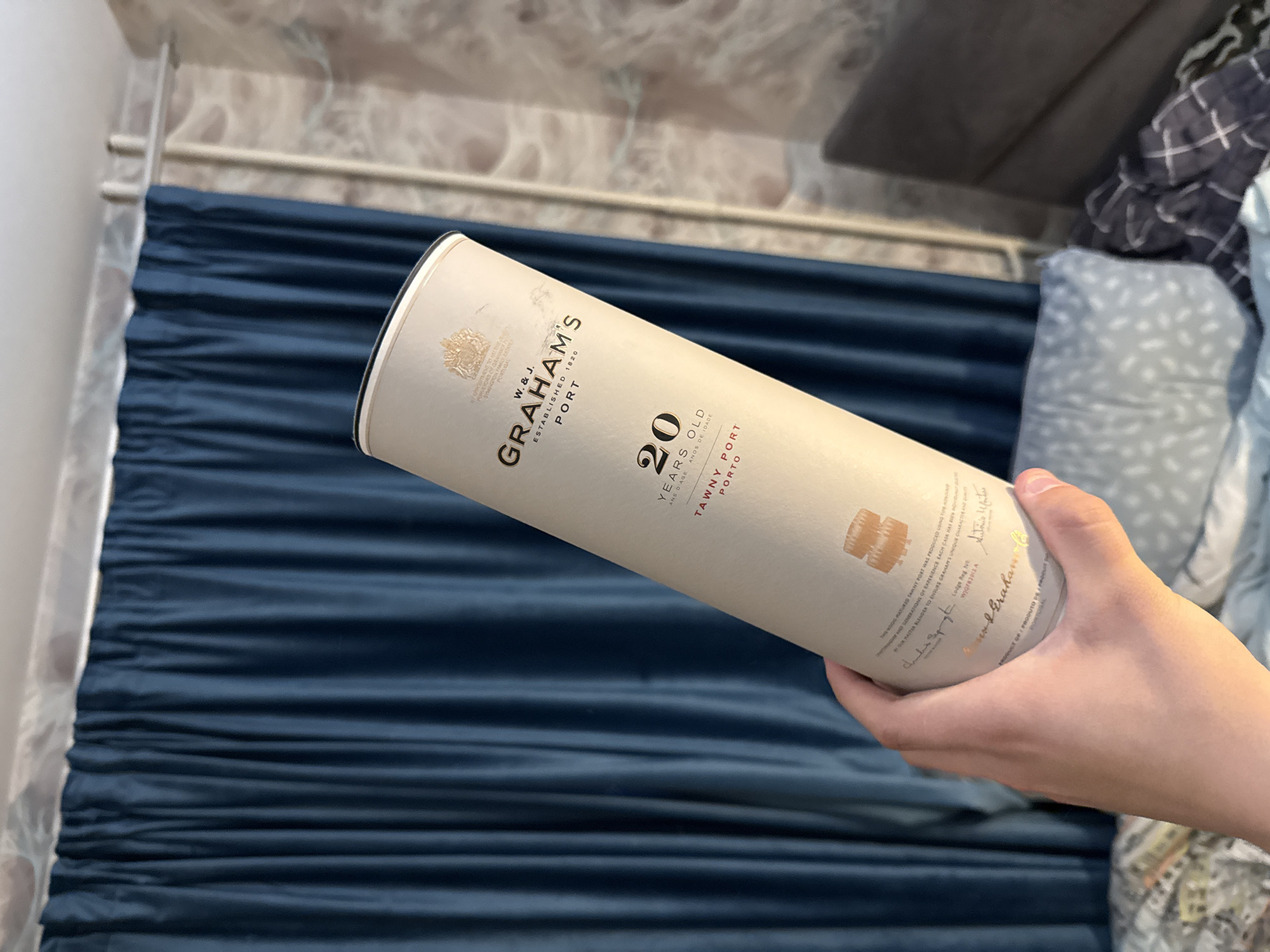 A hand holds a cylindrical tube of Graham's 20 Years Old Tawny Port. The tube is a light tan color with black text and gold accents. The text reads "W. & J. GRAHAM'S PORT ESTABLISHED 1820" near the top. Below that, it says "20 YEARS OLD" in large black numbers, followed by "TAWNY PORT" in smaller red text. The background consists of dark blue curtains and a patterned wallpaper. The image appears to be taken indoors.