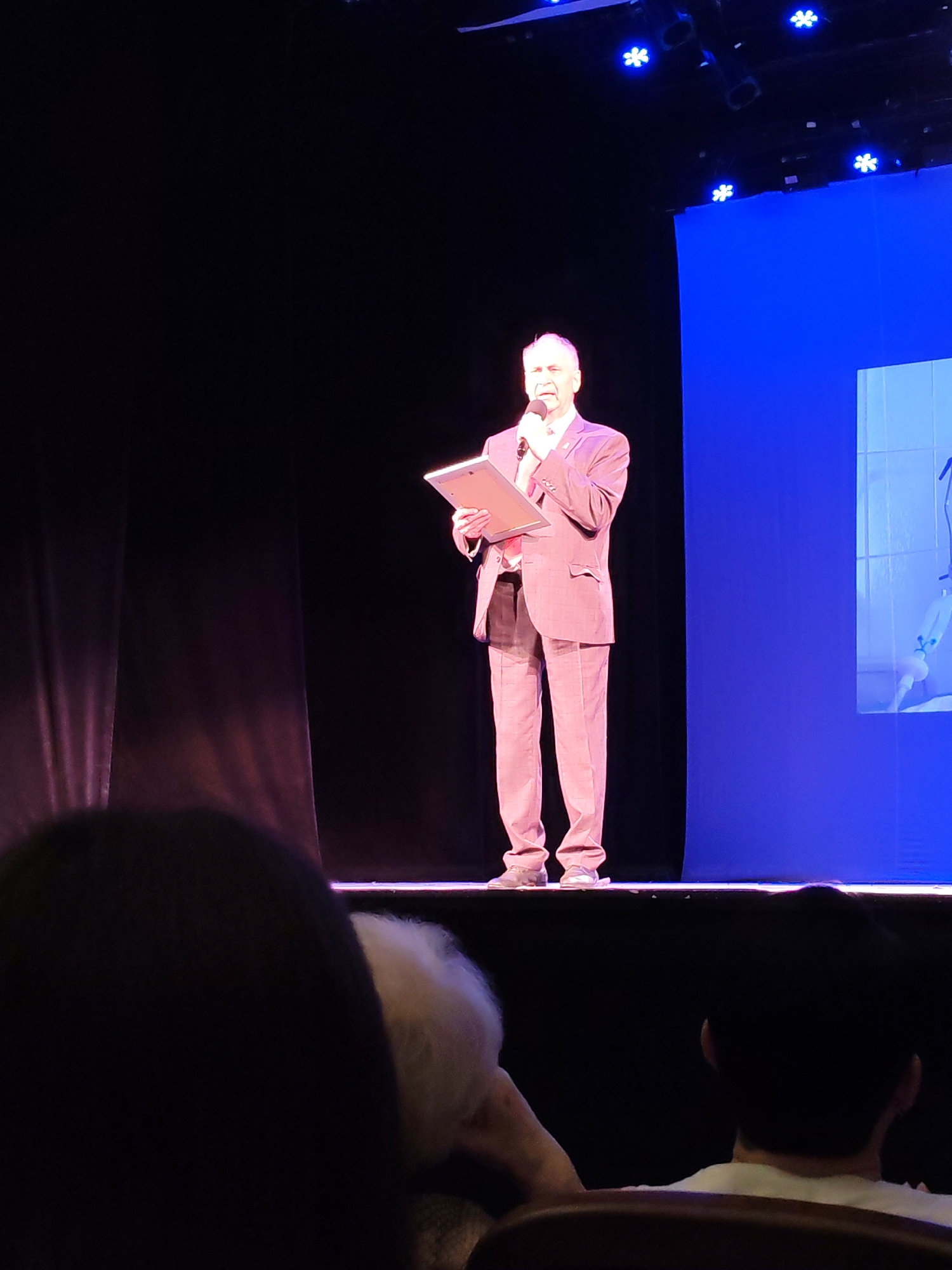 A man in a suit stands on a stage, holding a tablet and speaking into a microphone. He is illuminated by stage lights, while the background is dark with a large blue screen on the right. The audience is visible in the foreground, mostly out of focus. The image appears to be taken at a public event or performance.