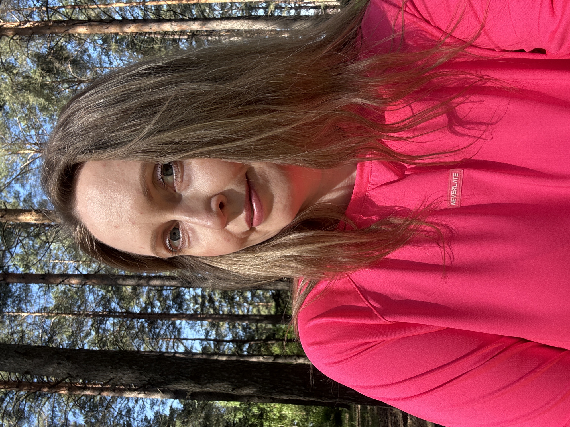 A woman with fair skin and blonde hair, wearing a bright pink top with the word "NEVERLATE" visible on the collar, is outdoors, likely in a forested area. Tall trees are visible in the background, with dappled sunlight filtering through the branches. The scene is potentially in Chaikovsky, Russia, based on the prompt's request.