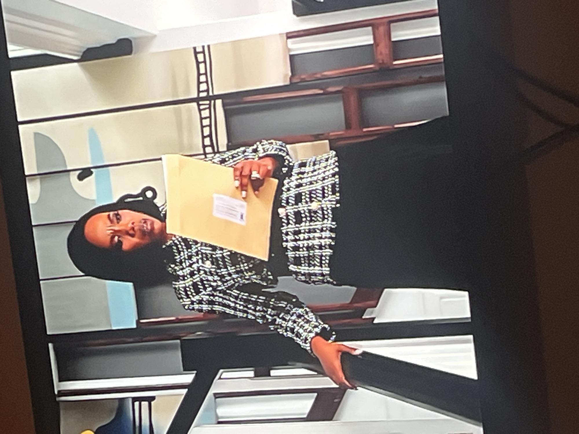 A woman, likely of African descent, is captured in a medium shot, leaning forward with a serious expression. She is wearing a black and white patterned jacket over a black outfit and is holding a light brown envelope. A ring is visible on her left hand. The background features a modern, abstract wall with geometric shapes in shades of grey, blue, and beige. Behind this, there are wooden shelves filled with white objects, possibly books or decorative items. The overall impression is that of an indoor setting, possibly an office or a reception area, with contemporary decor. The lighting appears to be artificial, and the time of day is indeterminate. The woman's gaze is directed towards the viewer, and her facial expression suggests concentration or perhaps a moment of contemplation. There is no visible text in the image.