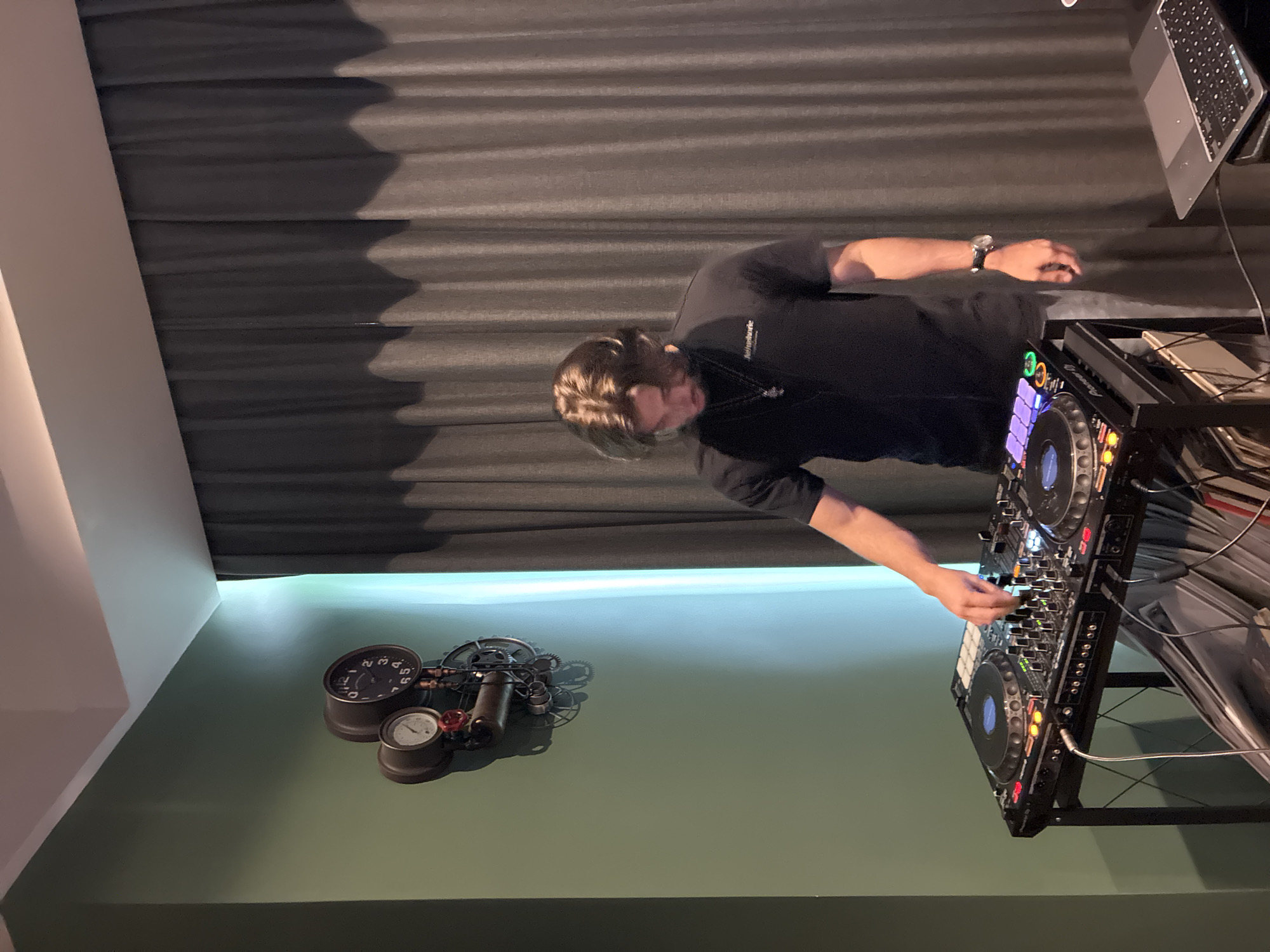 In an indoor setting within Huixquilucan de Degollado, Mexico, a male individual with medium-length brown hair, wearing a black t-shirt, light-colored pants, a necklace with a pendant, and a watch on his left wrist, is operating a black Pioneer DJ controller. The DJ controller rests on a black metal stand, displaying illuminated buttons and blue jog wheel centers. His right hand is positioned over the mixer section of the equipment. To the immediate right of the DJ setup, a silver laptop with an illuminated keyboard is open on a dark surface. The wall to the left is painted a muted green/teal color. Mounted on this wall, towards the upper left, is an ornate steampunk-style clock and gauge assembly featuring gears, pipes, and two circular gauges. The right side of the room is covered by full-height, pleated curtains that are dark gray at the top and lighter gray/beige at the bottom. A bright vertical light strip is visible between the green wall and the curtains. The ceiling is white.
