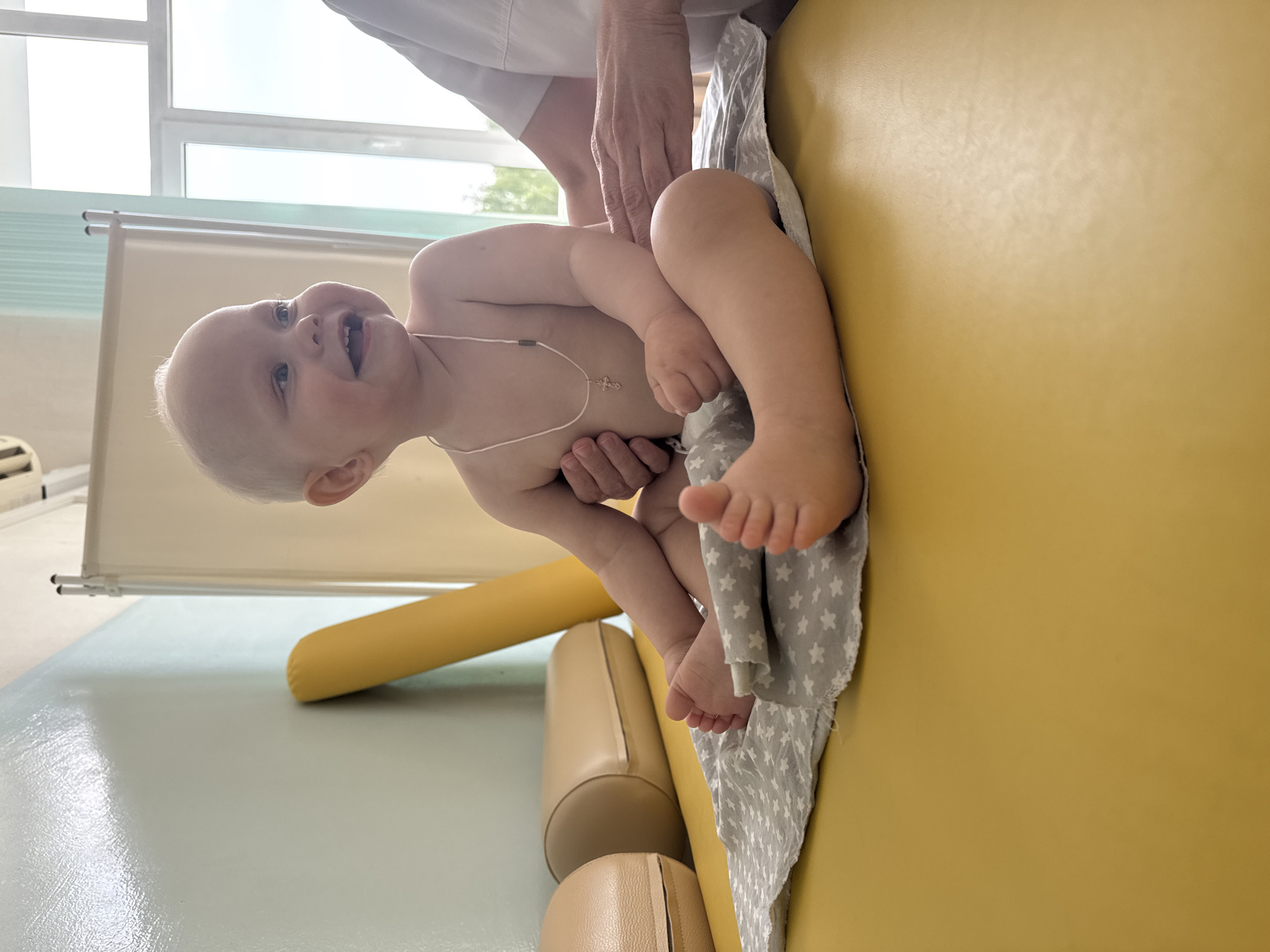 A baby with fair skin and blond hair is held up by an adult in what appears to be a medical or therapy setting. The baby is smiling and wearing a necklace with a cross pendant. The adult is wearing a white lab coat. The setting appears to be a room with a yellow padded table covered with a gray and white star-patterned cloth. There are other beige padded supports on the floor near the table. A window is visible in the background, as well as a beige privacy screen. The baby is located in Chaikovsky, Russia.