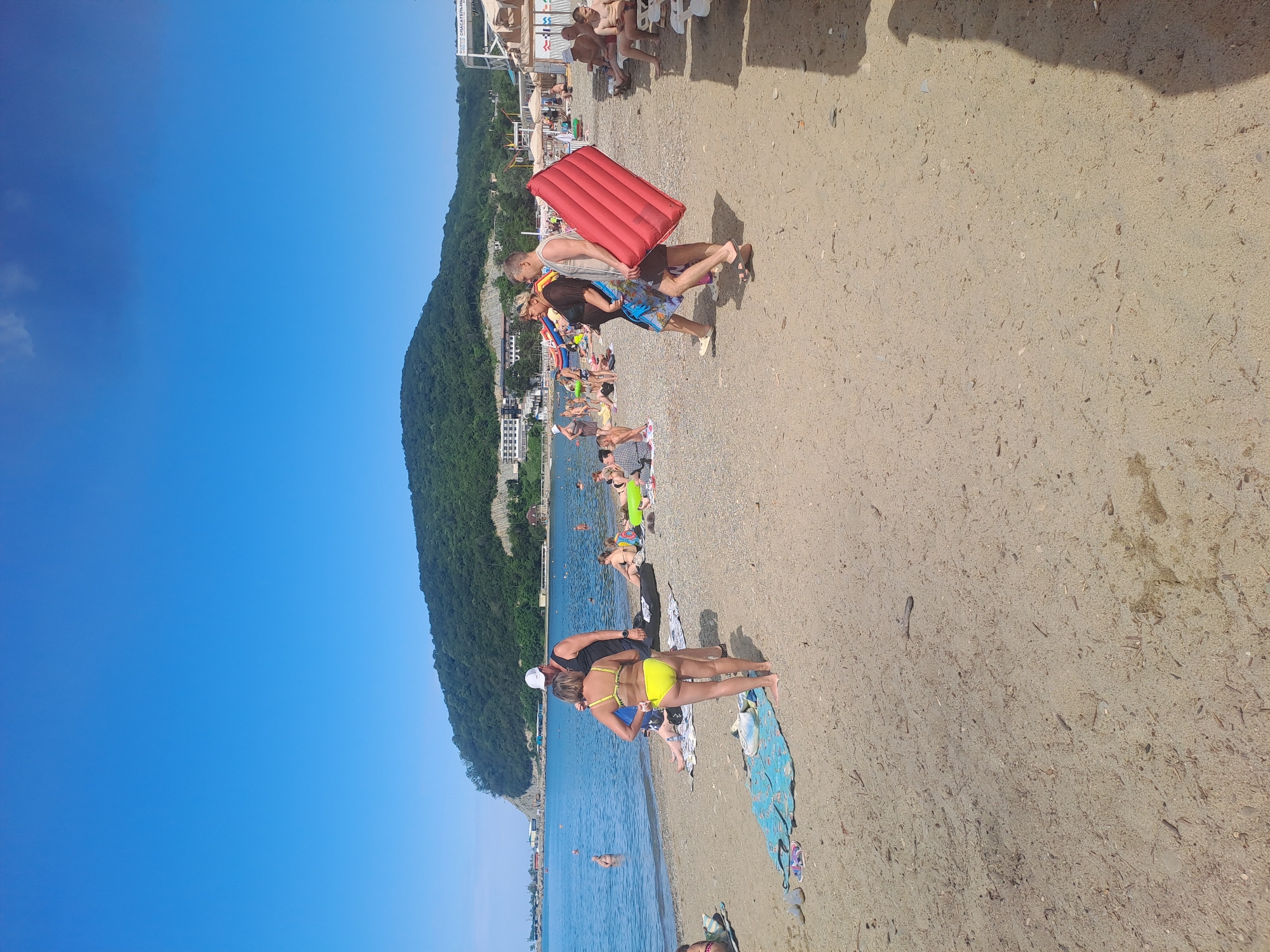 This image depicts a crowded beach scene in Dzhubga, Russia. The foreground is dominated by a sandy beach with scattered pebbles and small pieces of driftwood. Several beach towels and flip-flops are visible. In the middle ground, a group of people are interacting. A woman in a yellow bikini stands facing away from the camera, while a man in a black tank top and shorts stands beside her, talking. Further to the right, a couple walks along the beach, with the man carrying a large, deflated red inflatable mattress. Other beachgoers are scattered throughout the scene, some sunbathing, some in the shallow water, and some walking. The background features the calm blue sea, with a lush green forested hill rising behind the beach. Buildings and some infrastructure, possibly hotels or beach facilities, are visible on the hillside. The sky is clear and blue.