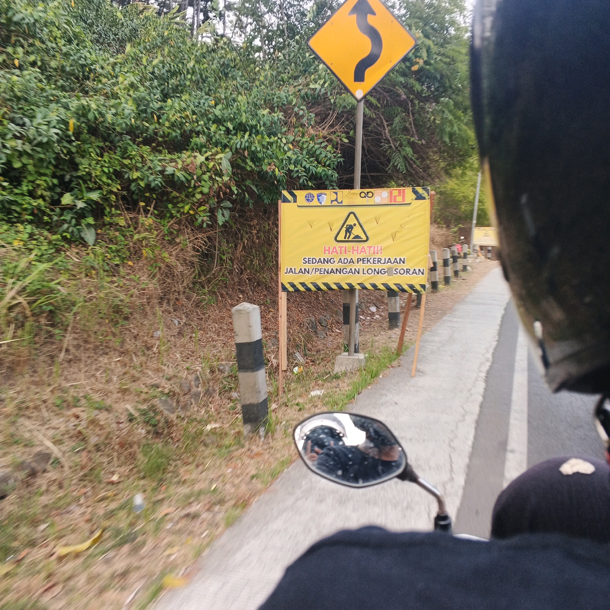 The image captures a roadside scene in Kebasen, Indonesia, where a motorcycle rider is passing a warning sign indicating road work. The sign, prominently displayed in yellow with black lettering and a pictogram of a person digging, reads "HATI-HATI!! SEDANG ADA PEKERJAAN JALAN/PENANGAN LONG SORAN," which translates to "BE CAREFUL!! ROAD WORK IS IN PROGRESS/SLIPPAGE HANDLING." To the left of the sign, a diamond-shaped yellow warning sign with a black curvy arrow indicates a winding road ahead. The background consists of lush green vegetation and trees lining the side of a paved road. The perspective is from the rider's point of view, with a partial view of their helmet and a motorcycle side mirror reflecting the surroundings. Several concrete posts with black and white stripes are strategically placed along the road. In the distance, another similar warning sign is visible, suggesting ongoing road maintenance or repair in the area. The overall scene depicts a typical Indonesian rural or semi-urban road environment during daylight hours.