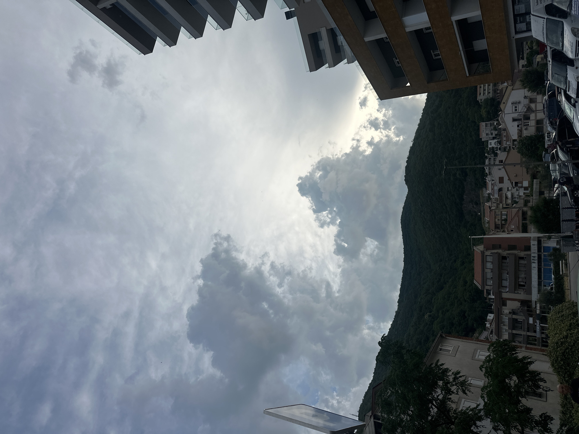 A street-level perspective captures a scene in Budva, Montenegro, with a dramatic sky filled with large, grey, and white clouds. The clouds are particularly dense and textured in the upper left and center of the image, with patches of brighter sky visible towards the right. In the foreground, modern apartment buildings with multiple balconies and glass railings dominate the right side of the frame. These structures are characterized by their clean lines and contemporary design, with one building featuring a warm, yellowish-brown facade. To the left, older, multi-story buildings with visible windows and shutters line a street. A lush, dark green, tree-covered mountainside forms the backdrop, rising steeply behind the buildings. In the lower portion of the image, a parking lot is filled with various cars, including vans and passenger vehicles. A few people are present, one walking in the parking lot and another partially visible in the lower left corner. A tall, white signpost with a reflective surface is positioned on the left side of the frame, obscuring a portion of the buildings and the sky. The overall impression is one of a city nestled against a natural, mountainous landscape under a dynamic, cloudy sky.
