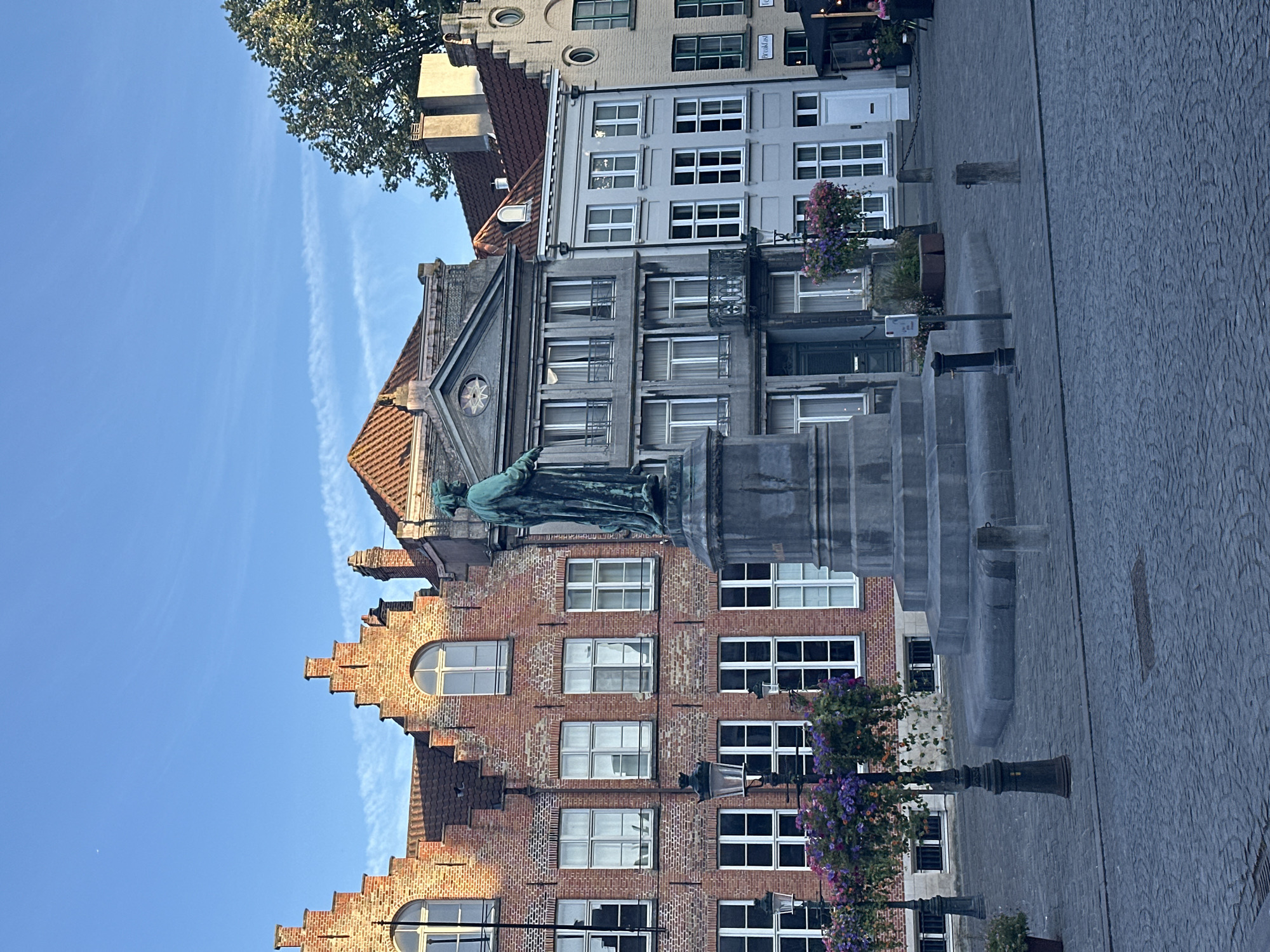 The image depicts a statue in a cobblestone square in Bruges, Belgium, with historic buildings forming the backdrop. The statue, cast in bronze, stands on a large, stepped stone pedestal at the center of the square. It portrays a figure in a long robe, facing to the right, with its left arm extended forward.
The surrounding buildings exhibit characteristic Flemish architecture. To the left, a red brick building features stepped gables and arched windows. Adjacent to it is a gray building with classical architectural elements, including pilasters and a decorative circular motif above the entrance. To the right, a pale yellow building with a mansard roof and dormer windows completes the row.
Streetlights with hanging baskets of purple and pink flowers are positioned in front of the buildings, adding a touch of color to the scene. The cobblestone pavement is worn, suggesting historical significance. The sky is clear with a few wispy clouds. The overall atmosphere is tranquil, suggesting an early morning or late afternoon setting due to the soft lighting. No people are visible in the frame.