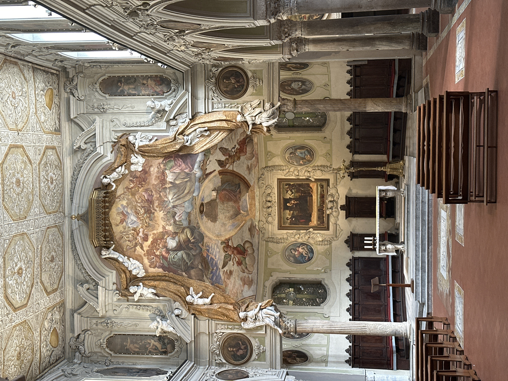 This image shows the interior of a church in Naples, Italy, specifically the Basilica of San Giorgio Maggiore. The view is towards the altar, with a large fresco dominating the wall above it. The fresco depicts a celestial scene with multiple figures, including what appears to be Christ in the center, surrounded by angels and saints.
Flanking the fresco are sculpted angels and ornate drapery. Smaller oval paintings are mounted on the walls on either side, depicting religious figures. The architecture features high ceilings with intricate decorative plasterwork and segmented panels. Large columns with Corinthian capitals support the arches of the side aisles.
In the foreground, wooden pews are arranged in rows on a reddish-brown tiled floor. The altar area is elevated, with steps leading up to it. The altar itself is covered by a white cloth and is flanked by decorative statues. Behind the altar is a large wooden choir stall. The overall impression is one of grandeur and religious solemnity.