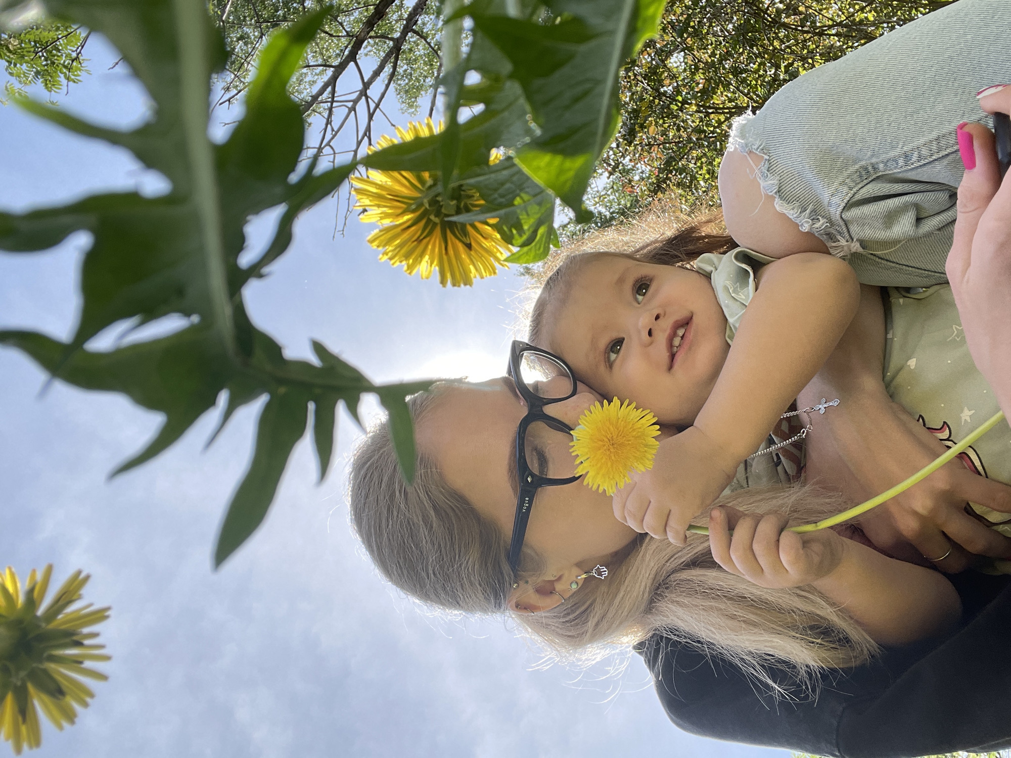 From a low angle, a fair-skinned adult with blond hair and black-framed glasses holds a young child. The adult is wearing earrings and is clad in a black garment. The child, with light skin and brown hair, wears a light green shirt with a pattern of unicorns. A dandelion flower is positioned between their faces. The adult appears to be kissing the child's cheek, and the child holds another dandelion. The sky is visible in the background, interspersed with the leaves and flowers of dandelions. The image was not taken in Izhevsk, Russia.