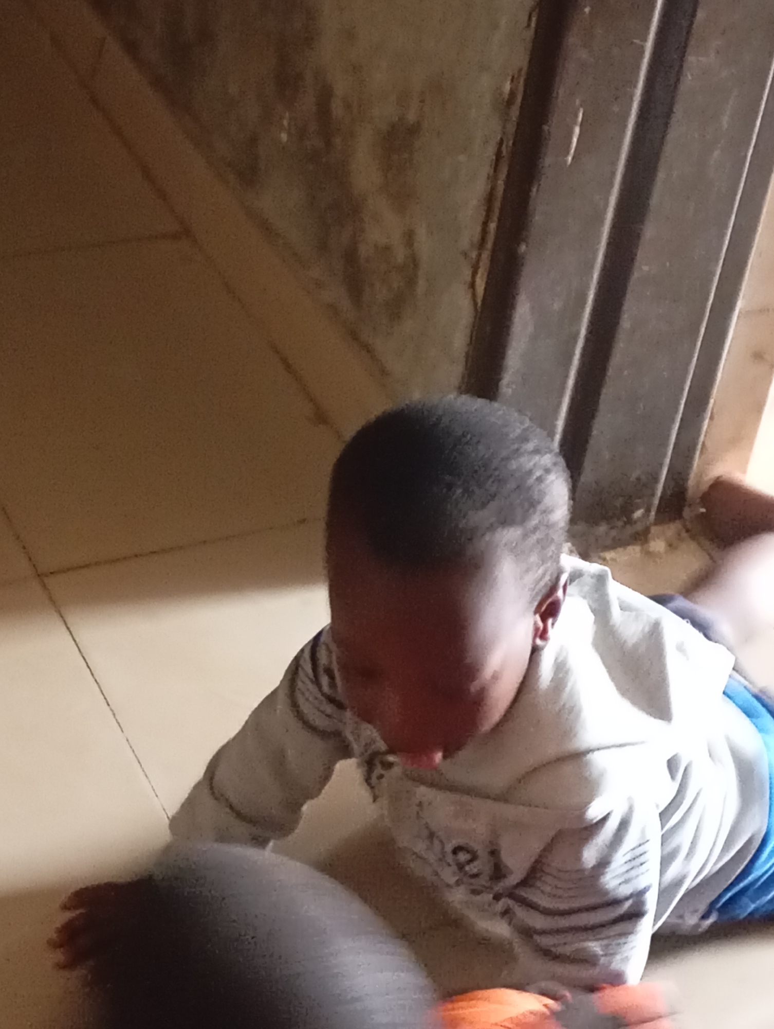 A young child is on the floor in a room. The child is wearing a gray hooded sweatshirt with a white lining and blue shorts. The child is on their hands and knees, with their head down, facing towards the floor. The floor appears to be tiled in a light brown color. To the right of the child, there is a dark metal door frame with vertical slats. The wall to the left of the child is a textured, off-white color with some dark stains. In the foreground, another child's head and shoulder are visible, out of focus.