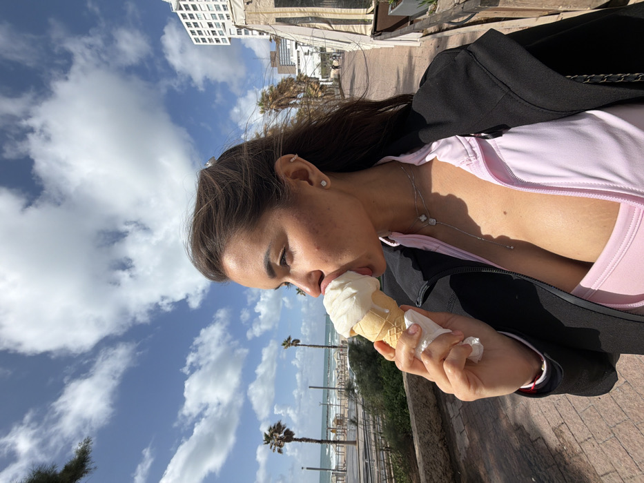 A young adult female is captured in profile, positioned off-center to the right, actively licking a large scoop of white ice cream from a waffle cone. She holds the cone in her right hand, which is wrapped in a white paper napkin. Her attire consists of a black zippered jacket worn open over a light pink top, revealing her décolletage. She has dark brown hair, which appears to be pulled back, and wears a silver necklace with multiple pendants, a small square stud earring, and a helix piercing in her visible ear.
The scene is an outdoor environment under a bright, partly cloudy sky, featuring white cumulus clouds against a blue background. The background reveals the Mediterranean Sea with moderate waves breaking towards a sandy beach. Several tall palm trees are visible between the subject and the sea. A paved promenade with a low stone wall borders the left side of the frame. In the mid-ground, distant human figures are visible on the beach or lower promenade. To the far right, a tall, multi-story white building with a distinctive architectural style is visible, alongside a partial view of a wooden or canvas awning-like structure closer to the foreground. This coastal setting, with its specific urban and natural elements, is consistent with the promenade and beach area of Tel-Aviv, Israel.