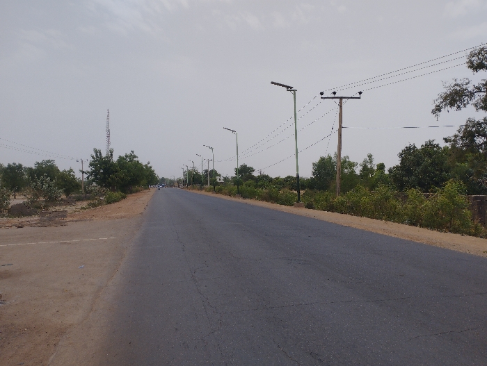 The image shows a paved road lined with solar powered streetlights and power lines in Kwadon, Nigeria. There are trees on both sides of the road, along with some dirt patches, suggesting a rural setting. The road is empty of vehicles and people, and the overall atmosphere is calm and quiet. The sky is a pale gray, suggesting a cloudy day.