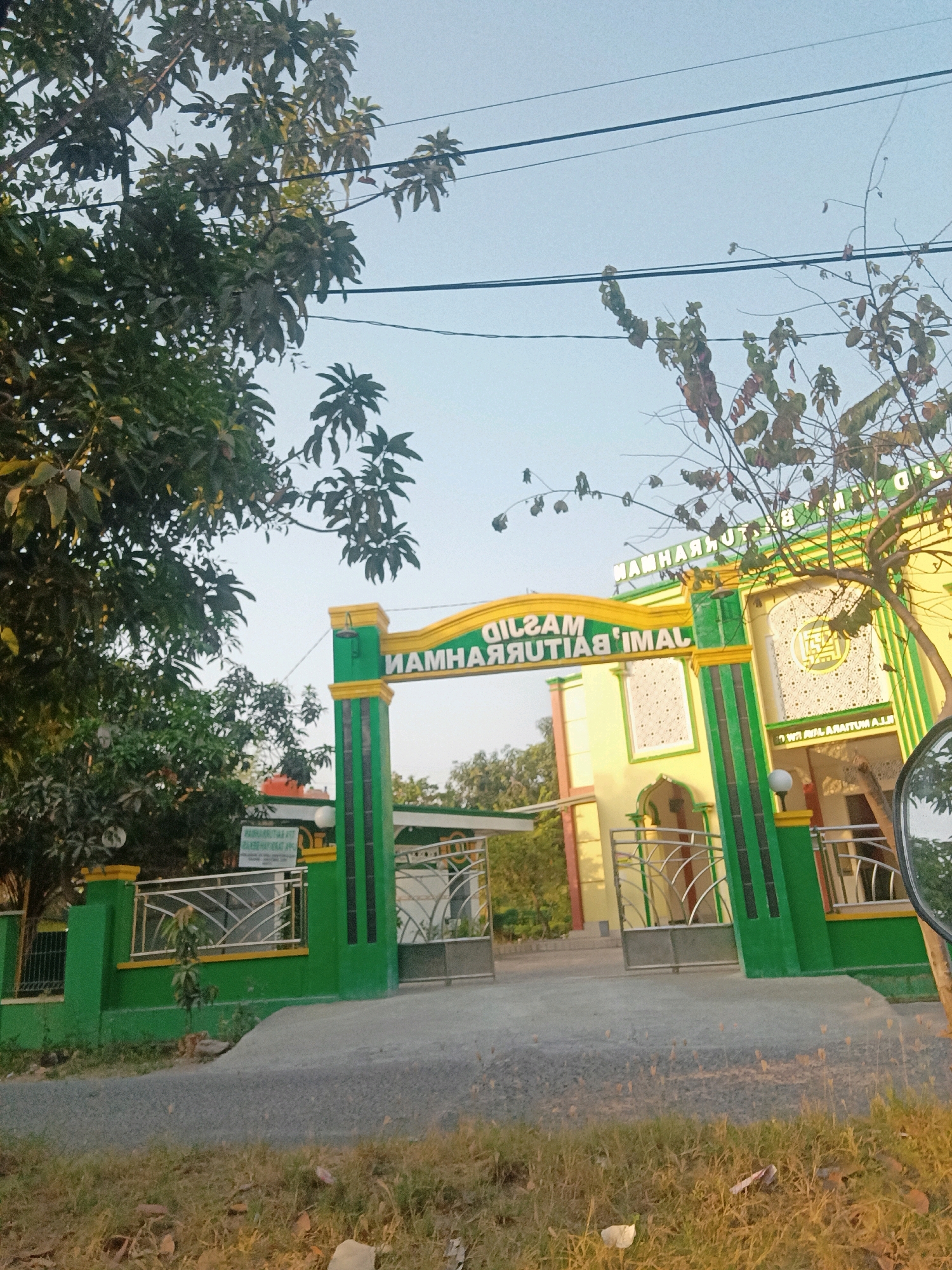 The image shows the entrance to a building identified by signage as "MASJID NURHIDAYAT" in Telagaasih, Indonesia. The architecture is characterized by a bright yellow facade with green pillars and trim around the entrance archway. The archway itself is adorned with yellow and green elements and features Arabic calligraphy that reads "MASJID NURHIDAYAT". In front of the building, there is a paved walkway leading to a gate. The gate has green railings with a decorative metal fence. The surrounding environment includes trees with lush green foliage, power lines overhead, and a clear sky. The ground in the foreground is covered with dry grass and some scattered leaves, suggesting a natural or semi-urban setting. The overall impression is of a religious building in a tropical or subtropical climate.