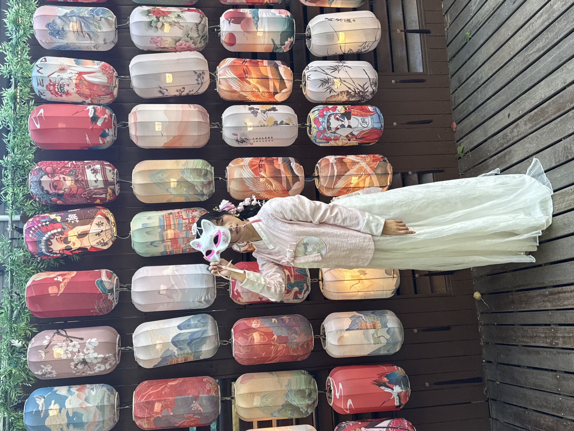A person wearing a light pink long-sleeved robe and a white dress, along with a white mask with pink highlights, is lying down in front of a wall covered with rows of colorful lanterns. The backdrop is a brown wooden structure, and the lanterns display a variety of designs, including floral patterns, landscapes, and traditional Chinese figures. The person is holding the mask up. The scene appears to be outdoors, with green foliage visible on the side of the lantern wall and a wooden deck on the side of the person. The location is Shangcheng District, China.
