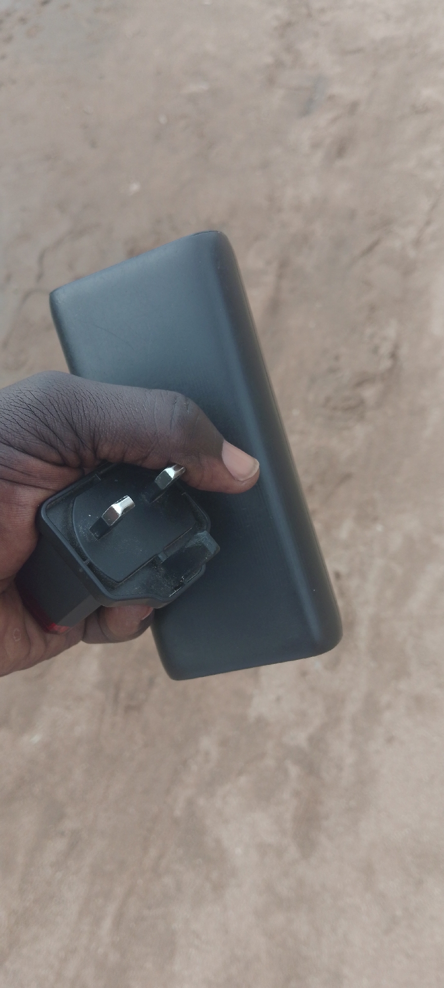 A person's dark-skinned hand is holding a black power bank and a black electrical plug. The power bank is rectangular with rounded edges. The electrical plug has three silver prongs. The background is a neutral, textured surface, likely the ground, with variations in color and small debris. The scene is outdoors in Jalingo, Nigeria.