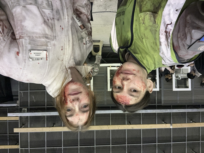 Two adult females are positioned in the foreground of an indoor scene, appearing to take a selfie. Both subjects exhibit simulated blood on their faces and clothing, indicative of special effects makeup. The female on the left has dark hair and wears a green high-visibility vest over a dark garment, both stained with simulated blood. A dark patch with a silver emblem is visible on her right shoulder area. The female on the right has lighter hair and wears a white full-body suit, extensively splattered with simulated blood. An identification badge with the name "EMMA JOHNSON" and a barcode is affixed to her upper chest. A red patch displaying "NEO" and numerals is visible on her left shoulder area. In the blurred background, several individuals are present, some engaged with mobile devices or cameras. The environment appears to be a studio or soundstage, characterized by large, dark, louvered wall panels, exposed horizontal wooden beams, and vertical metal supports. White rectangular frames and a table are visible in the mid-ground. The scene is located in Huangdao District, China.