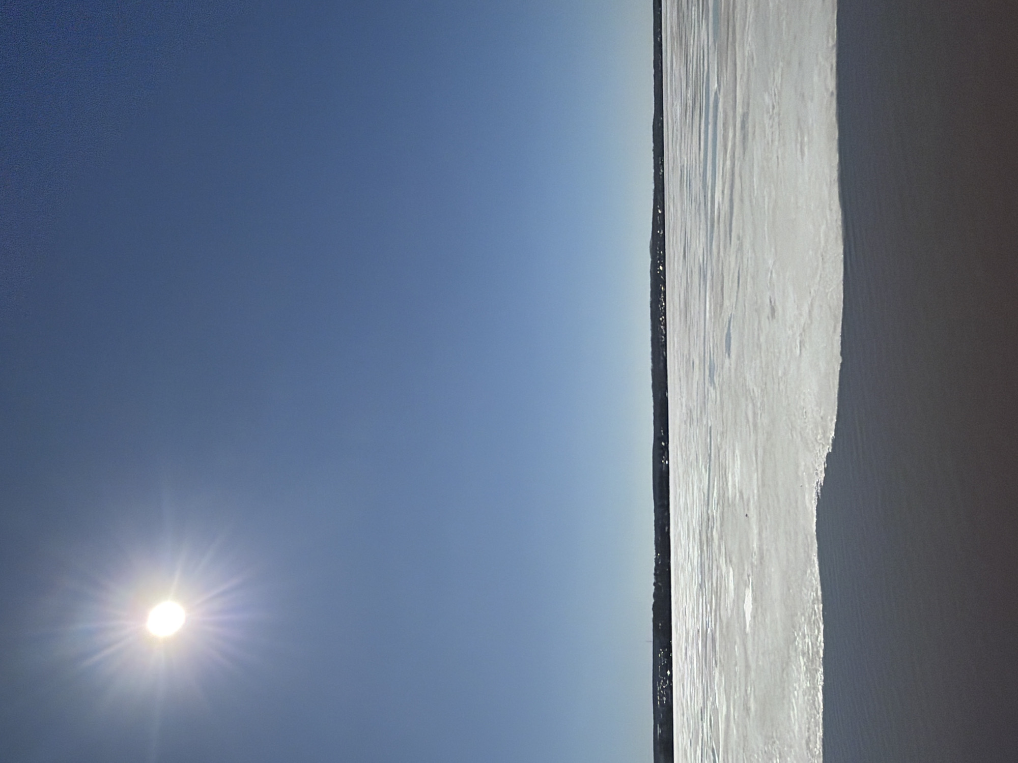 The image depicts a bright, sunny sky with a prominent sunburst. Adjacent to the sky is a body of water, possibly a lake or river, with reflections on its surface. A thin strip of land with dark vegetation separates the sky and water. Based on the mention of Votkinsk, Russia, and the type of landscape, it is plausible that this scene is of the Votkinsk Reservoir, which is a significant feature of the city and region. There are no people visible.