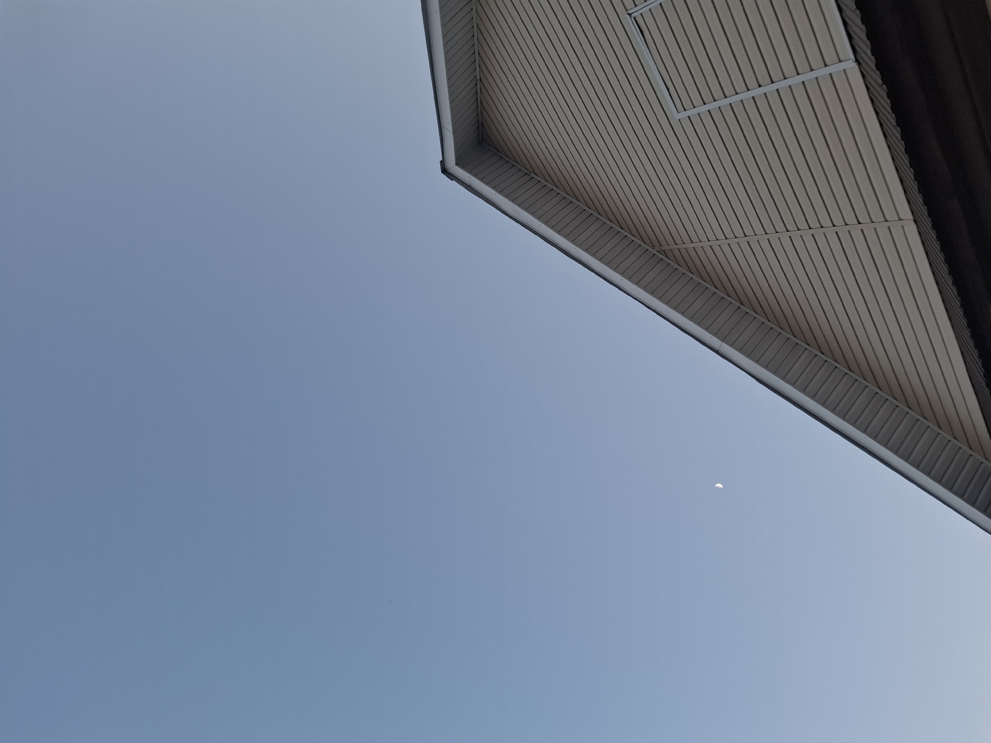 The image depicts a view looking up at the corner of a building's eaves against a clear, light blue sky. The eaves are composed of horizontally laid panels with a light brown or beige tone and are trimmed with a grey or silver-colored edge. A small, crescent moon is visible in the sky near the eaves. The location is Амзя, Russia.