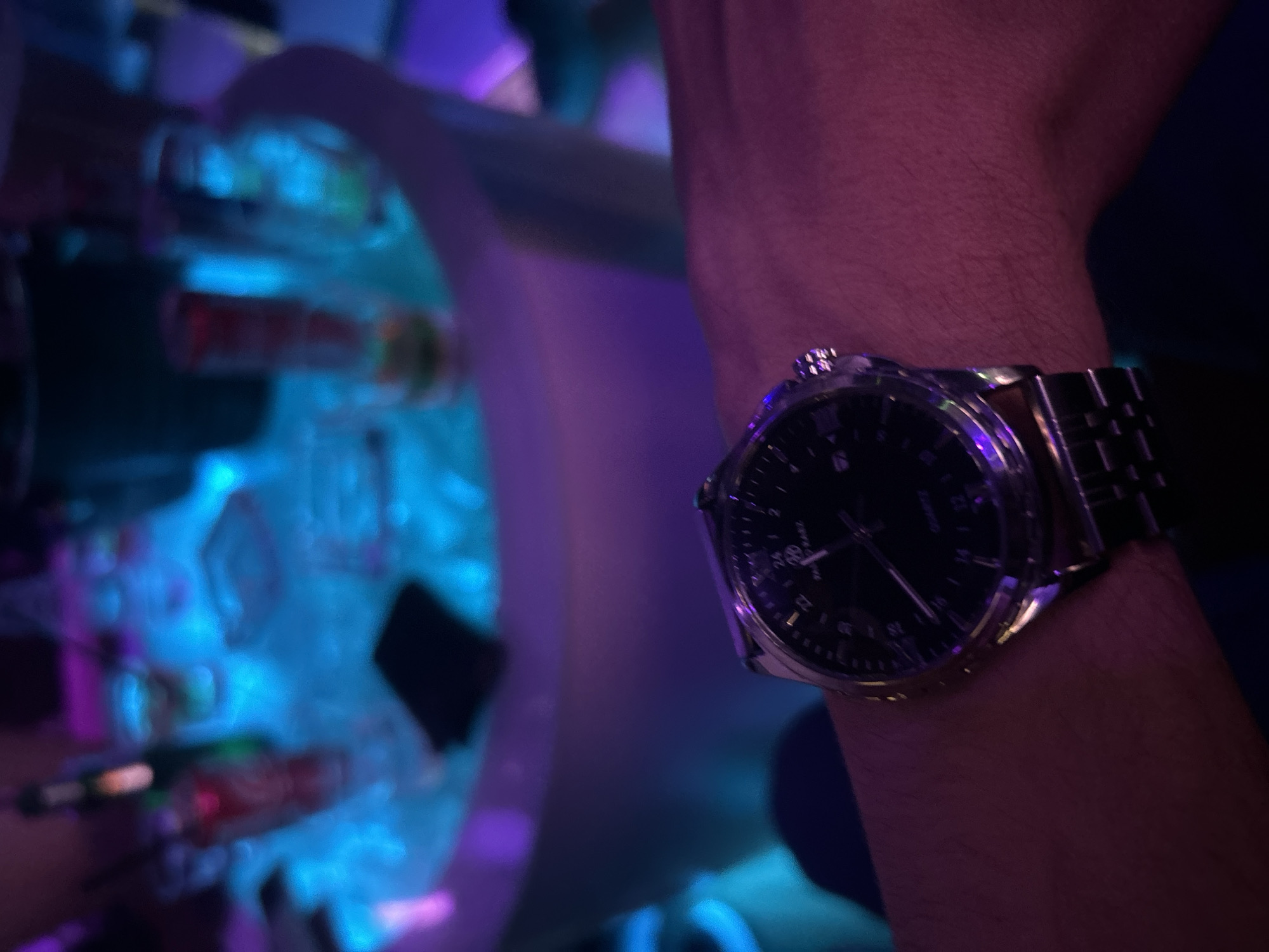 The image shows a wrist wearing a stainless steel watch. The background is blurry and features what appear to be test tubes or containers with colored liquids, illuminated by blue and purple lighting. It's difficult to determine the exact location, but the context suggests the image was taken in the city of Veliko Tarnovo, Bulgaria. It's impossible to ascertain any information of a forensic nature from this image.