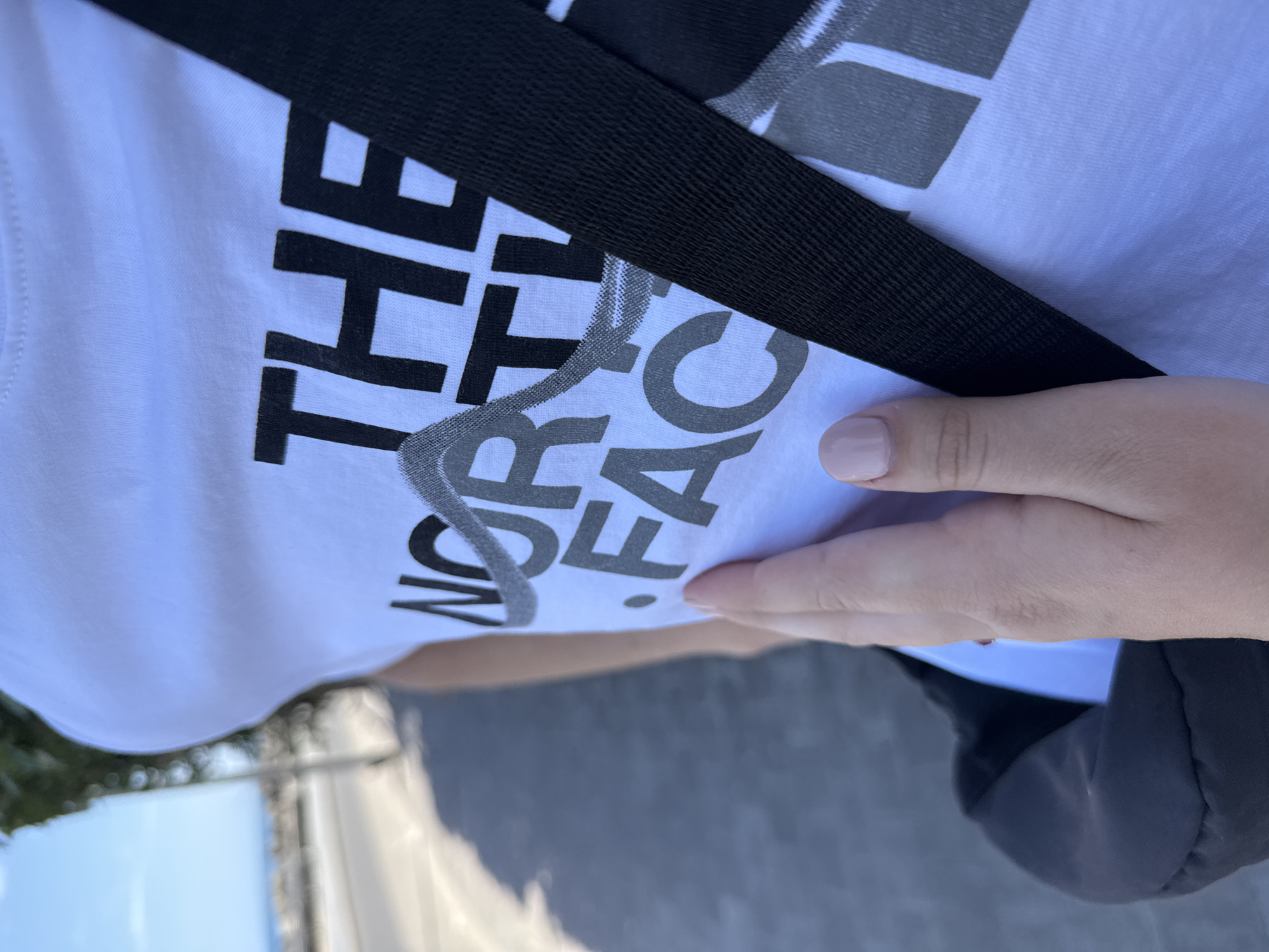 The image shows a person wearing a white t-shirt with "THE NORTH FACE" printed on it in black and grey. A black strap crosses the shirt diagonally. The person's hand is visible, with a light pink nail polish. The background shows a glimpse of a street or sidewalk, suggesting the location is outdoors.
