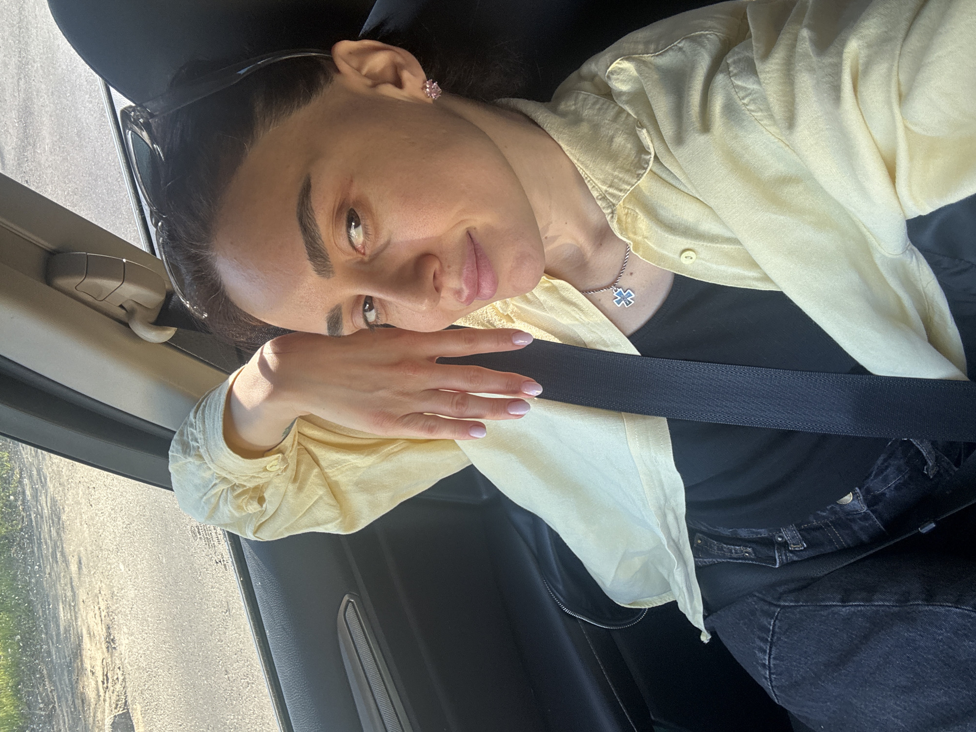 A woman with dark hair is sitting in a car, likely the passenger seat. She is wearing a light yellow shirt, a black t-shirt, and dark jeans. A seatbelt is across her chest. She has a necklace and earrings. Sunglasses are perched on her head. Her head is resting on her hand. The background shows an out-of-focus view of a road and trees, which suggests the location might be in the vicinity of Tbilisi, Georgia.