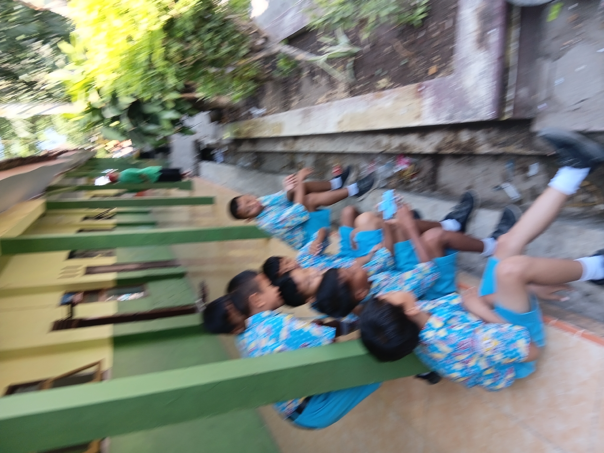 The image depicts a group of male students, likely elementary school age, sitting on a tiled ledge outside a building with a yellow and green facade. They are wearing matching blue and white patterned shirts and blue shorts or pants. Some students are looking down, while others are looking towards the camera or away from it. The environment appears to be outdoors, with a drainage ditch running alongside the ledge. Green foliage is visible in the background. The scene is consistent with a school setting in Tanggul, Indonesia, where such school uniforms and outdoor walkways are common.
